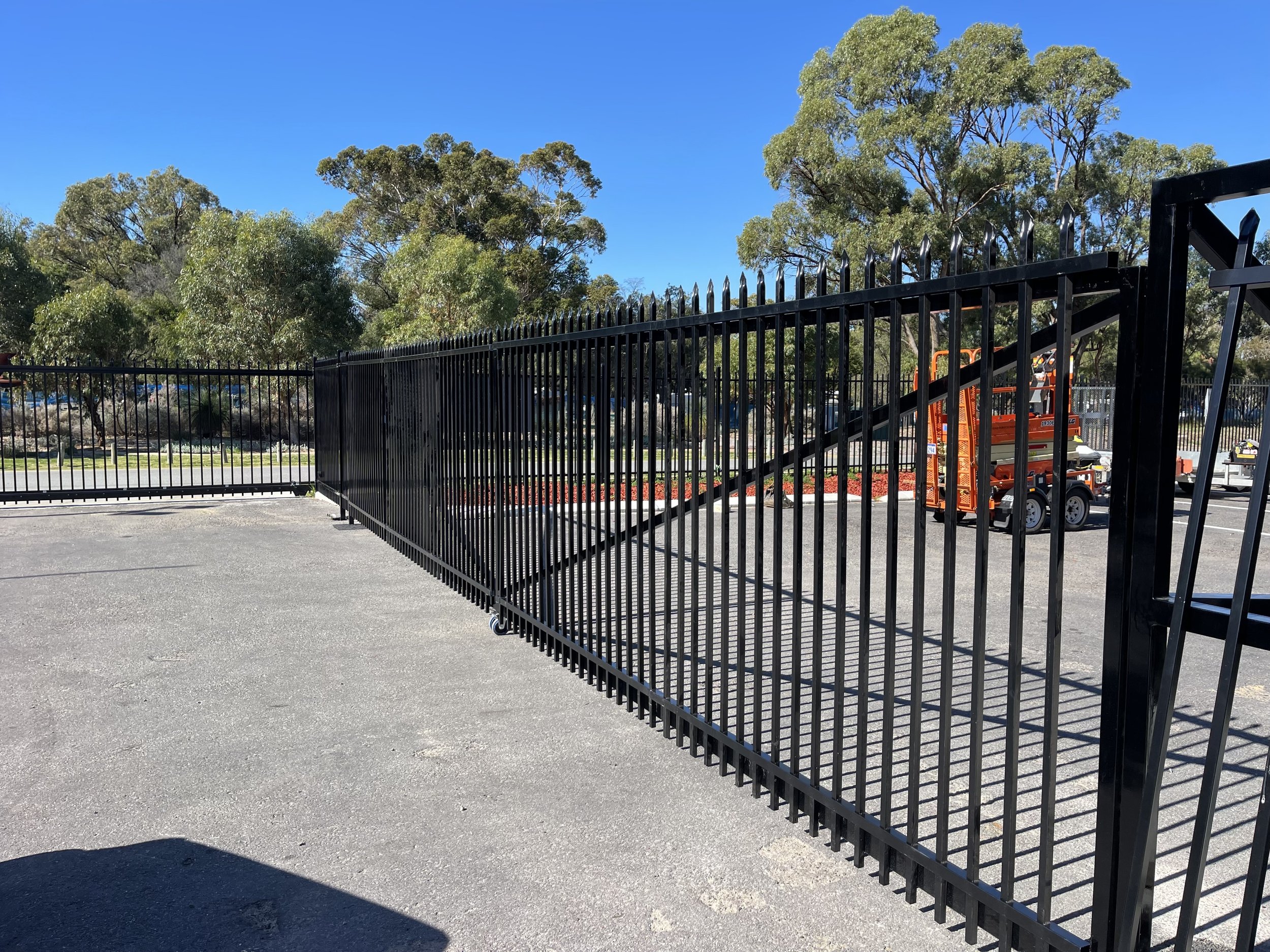 A black metal fence on a paved surface with a bright blue sky and trees in the background. There is orange construction equipment near the fence.
