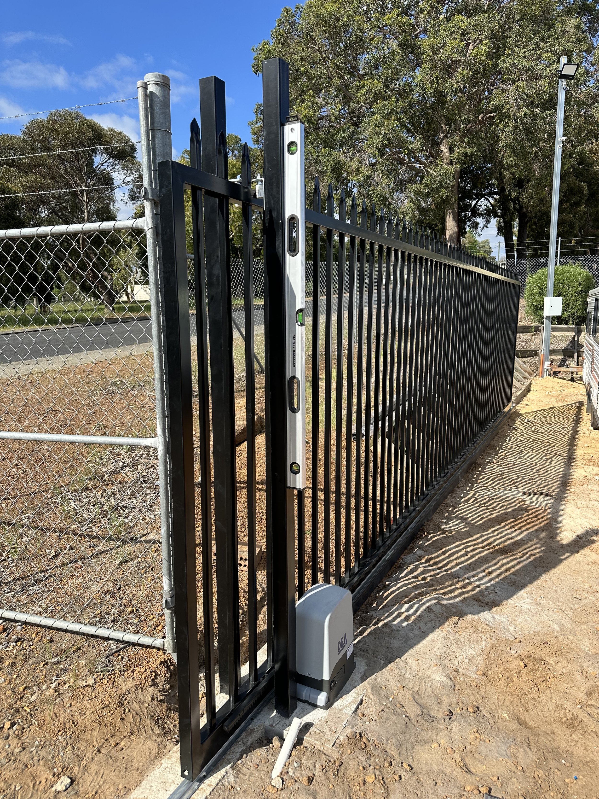 Black metal slide gate with a pedestrian electric lock system, installed on a dirt pathway next to a chain-link fence, under a clear blue sky with trees in the background.