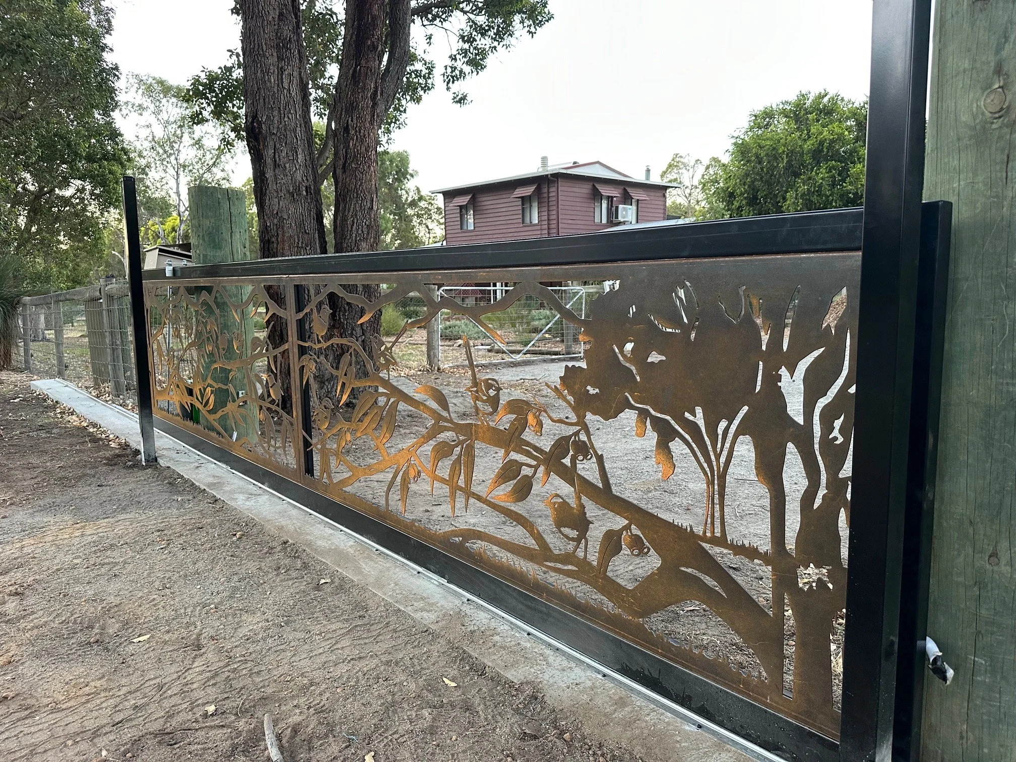Decorative metal gate with nature-inspired cut-out design, shown beside a dirt path in a park or zoo area.