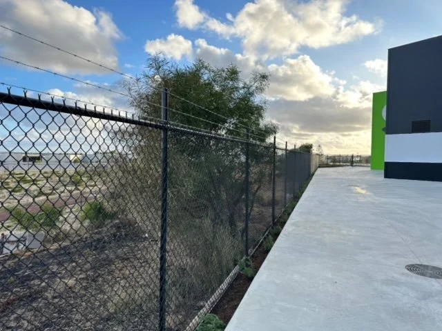 A chain-link fence separating a commercial building from a dry, grassy area with scattered bushes and trees under a partly cloudy sky.