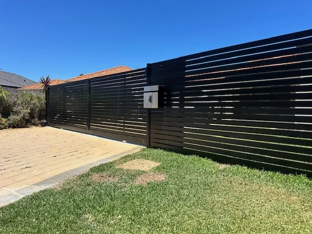 Black horizontal slat fence with mailbox labeled '4', adjacent to a paved walkway and a grassy lawn, under a clear blue sky.