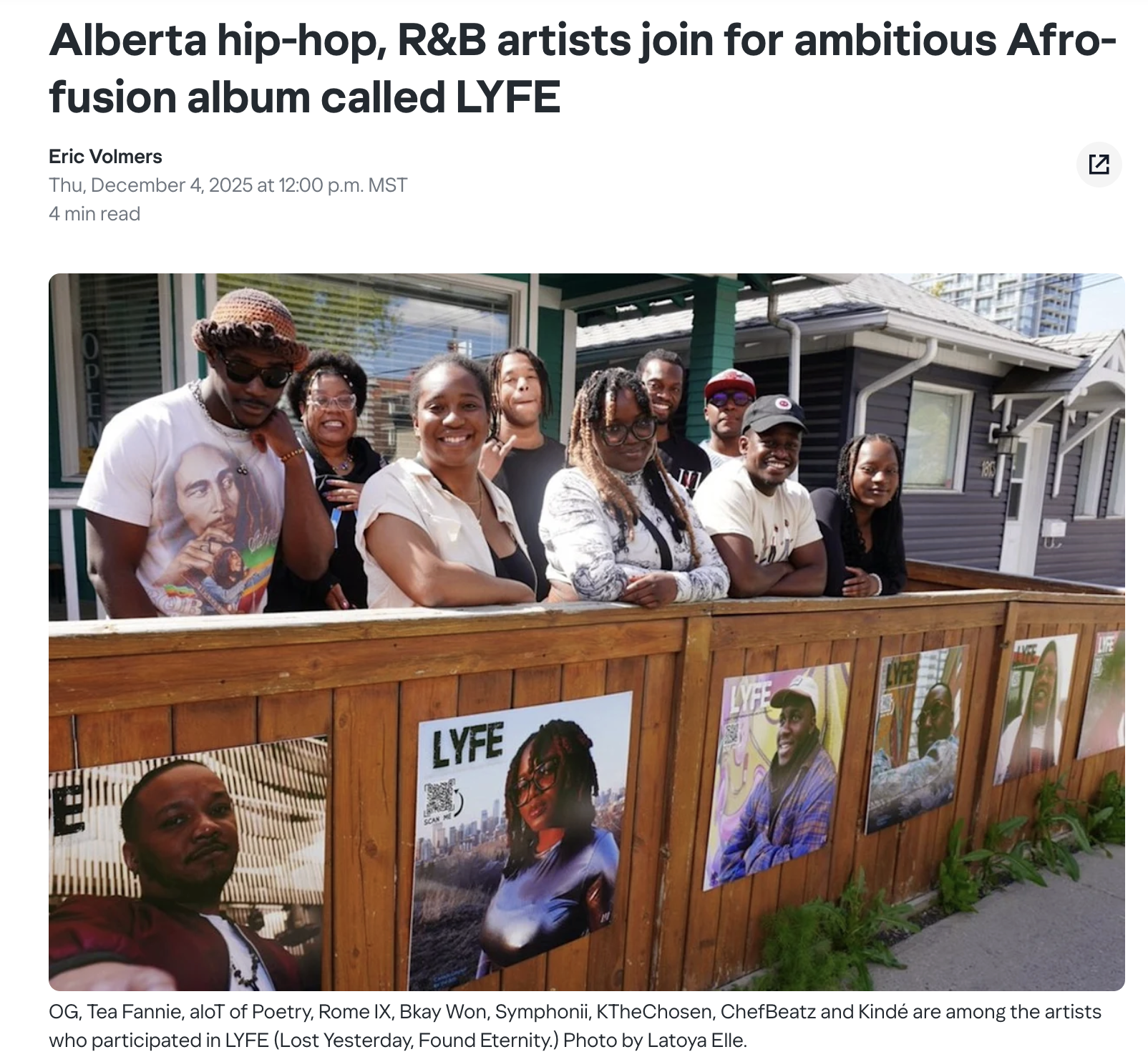 Group of people standing behind a wooden fence with posters of artists displayed on it, taken outdoors in front of a house with tall buildings in the background.