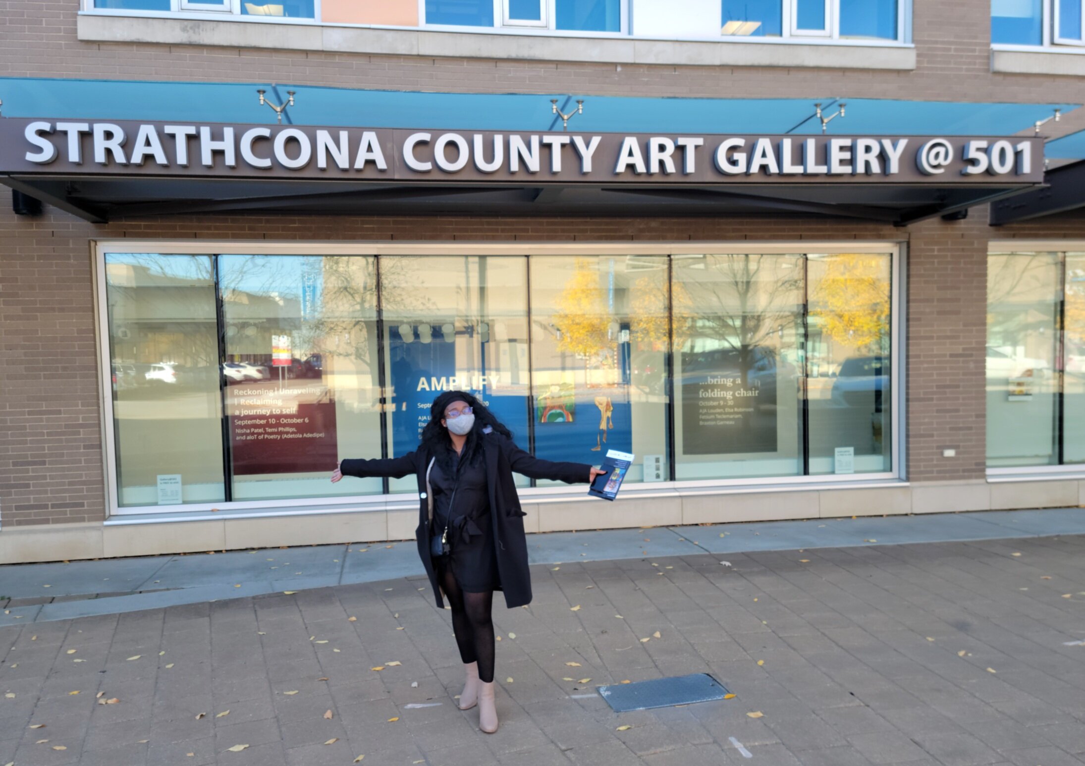 Person wearing a face mask standing outside the Strathcona County Art Gallery holding a pamphlet and extending their arms.