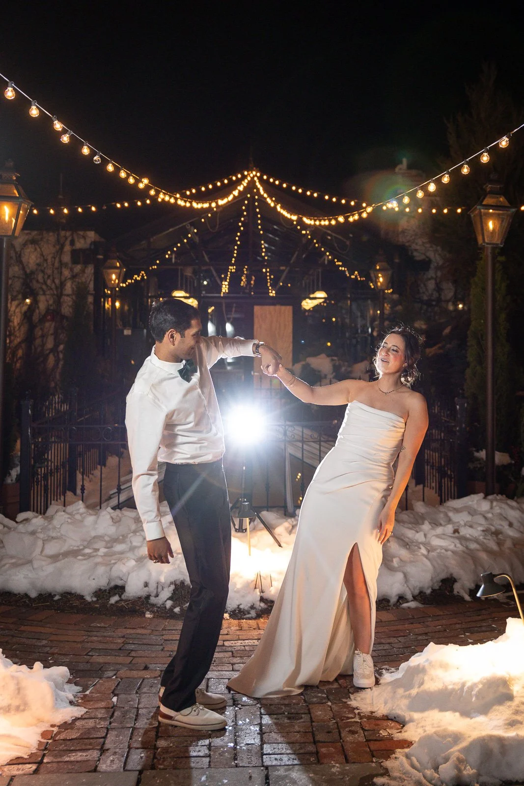 A couple dancing outdoors at night, dressed in formal attire, with snow on the ground and string lights overhead.