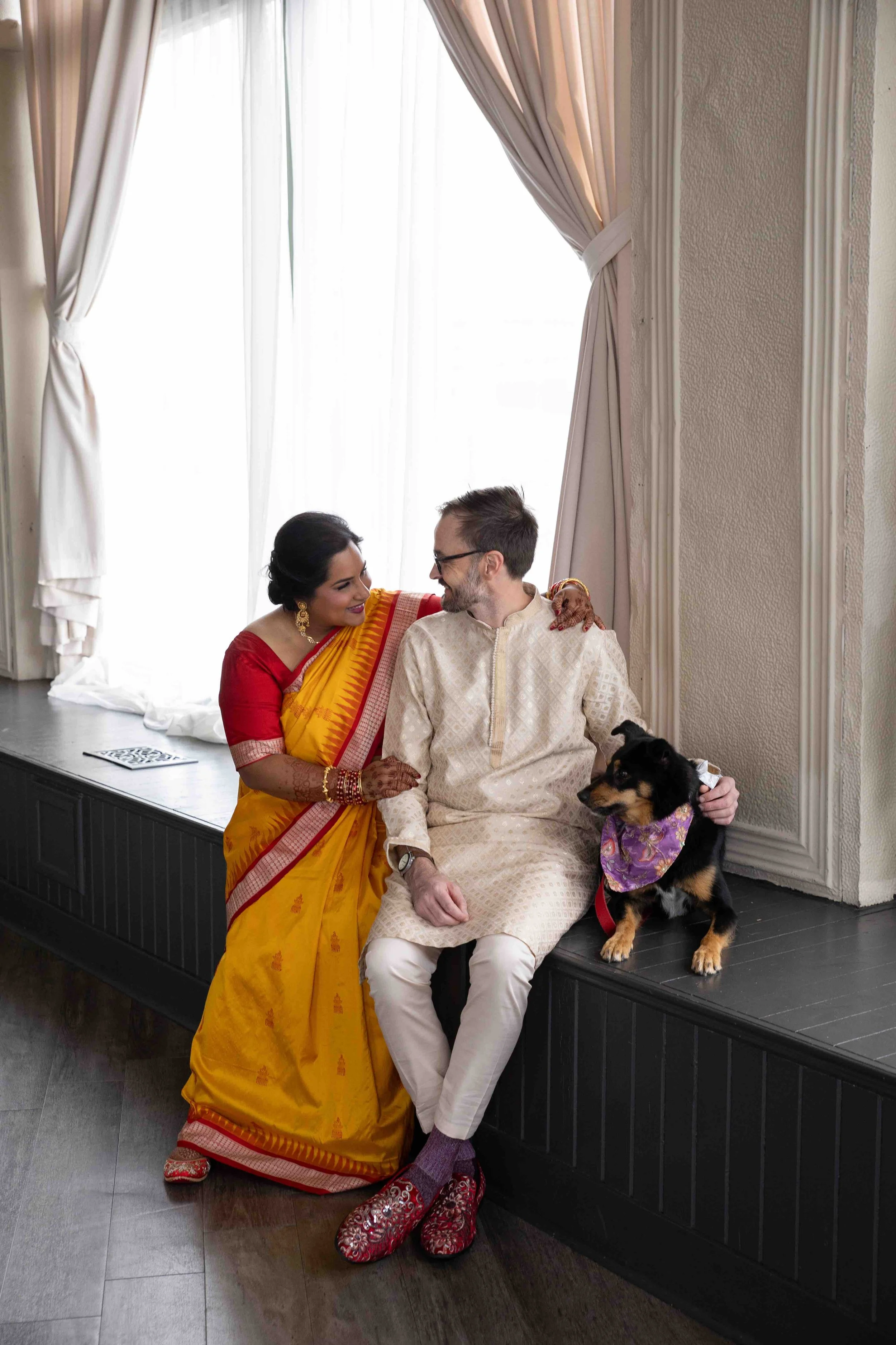 A couple in traditional Indian attire sitting on a window ledge with their dog. They are smiling at each other in a warmly lit room with curtains.