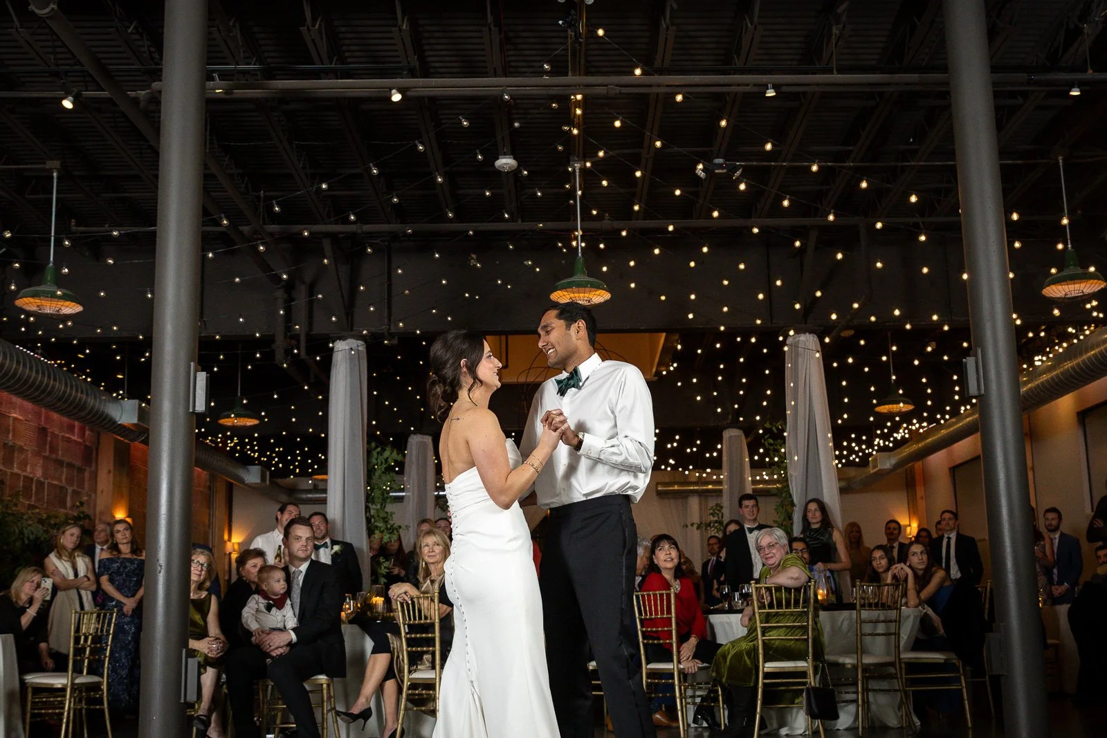 A bride and groom dancing at their wedding reception with guests watching, decorated with string lights and elegant decor.
