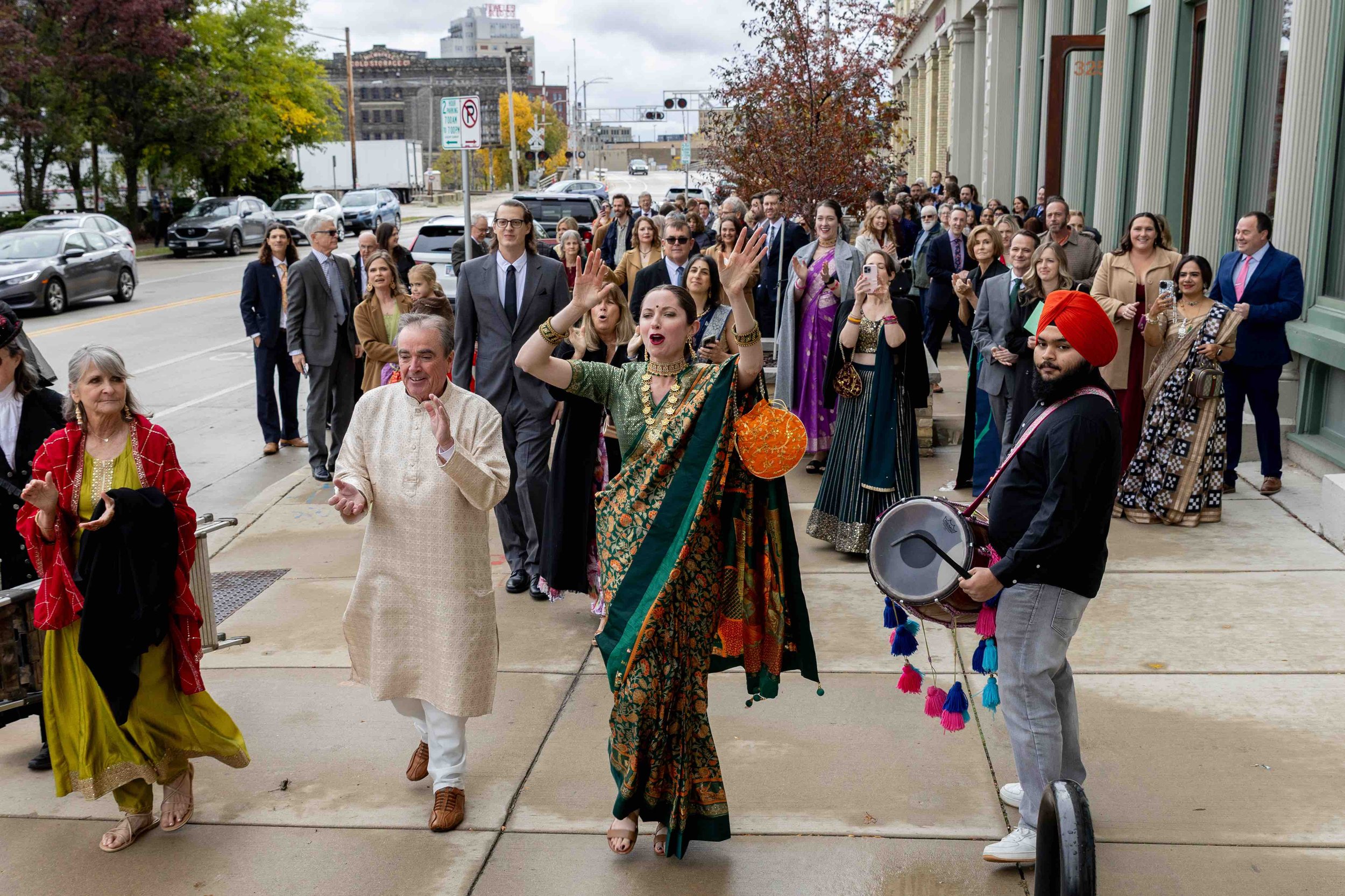 Group of people walking on a city sidewalk during a cultural celebration, with a woman in traditional Indian attire dancing and a man in a turban playing a drum.