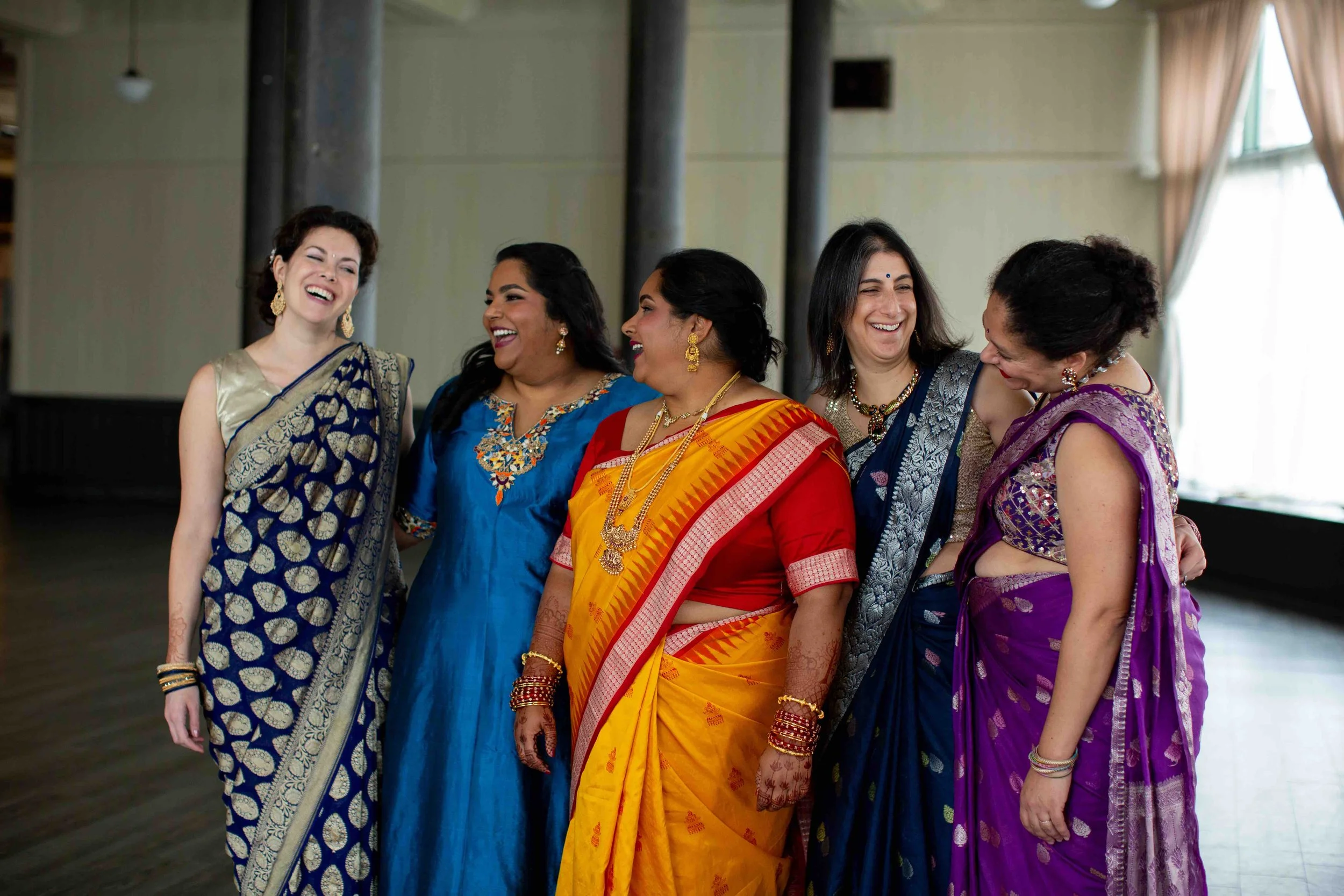 Five women dressed in colorful traditional Indian sarees are laughing and talking in a spacious room with large windows.