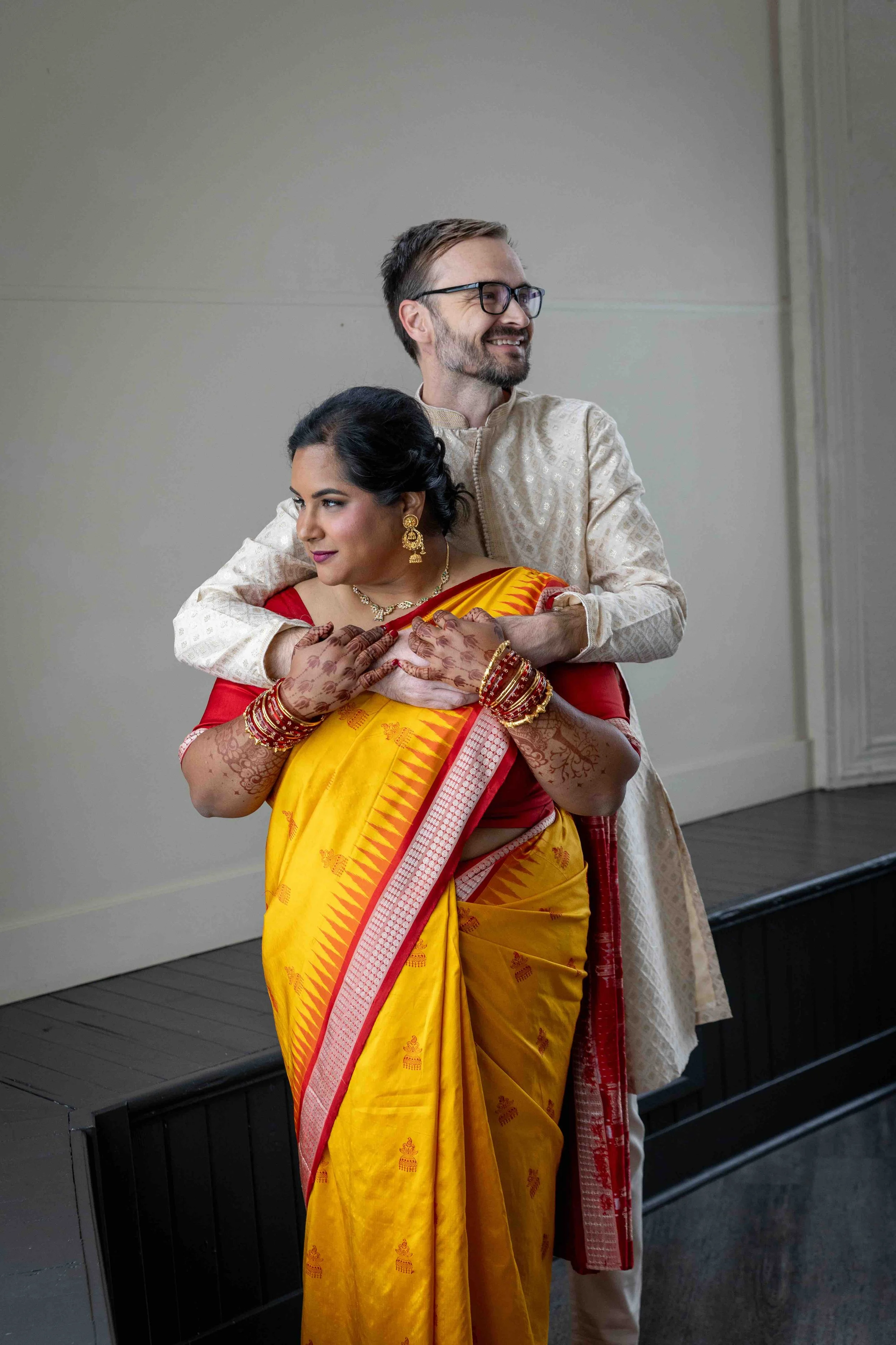 A couple, the woman in a yellow saree with red accents and the man in traditional cream attire, embracing during a celebration or ceremony.