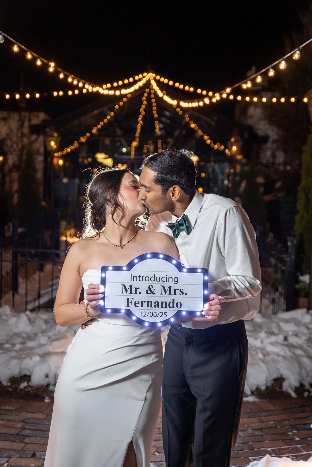 A couple shares a kiss at night; the woman holds a sign with their names and wedding date, illuminated by decorative lights.