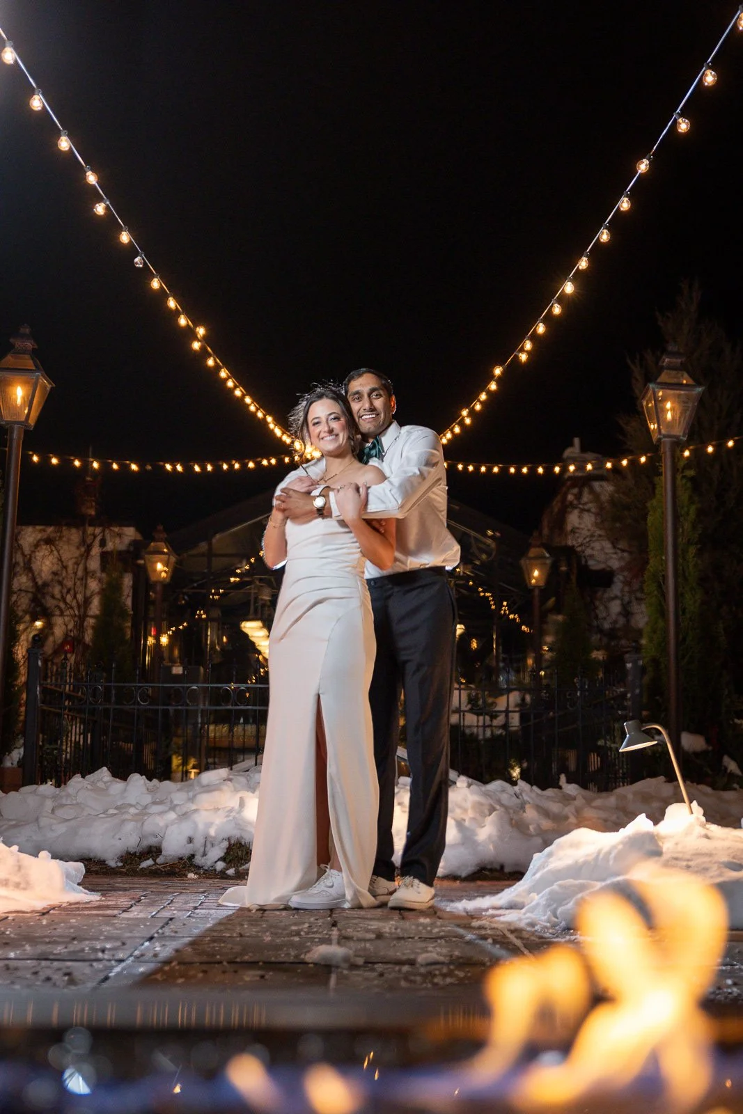 A couple dressed in formal attire, standing close and smiling, celebrating outdoors at night with string lights above and snow on the ground.
