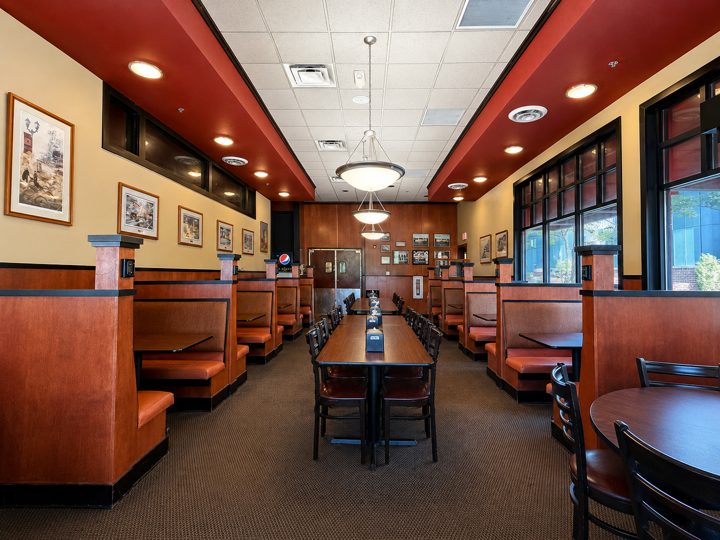 Empty restaurant with booth seating along the walls, a long central dining table with chairs, framed pictures on the walls, large windows on the right, and ceiling lights.