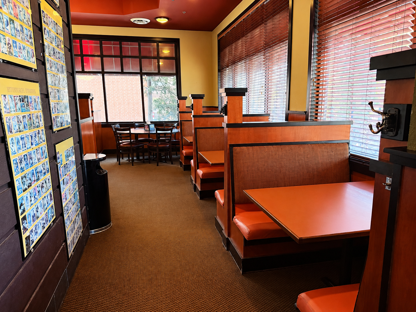 Empty booths with orange and brown upholstery inside a restaurant with large windows and blinds, a small table with chairs near the window, and a wall display of colorful coupons.