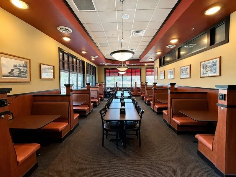 Empty restaurant dining area with booths along the walls, a central table with chairs, framed artwork on the walls, windows at the back, ceiling lights, and red window shades.