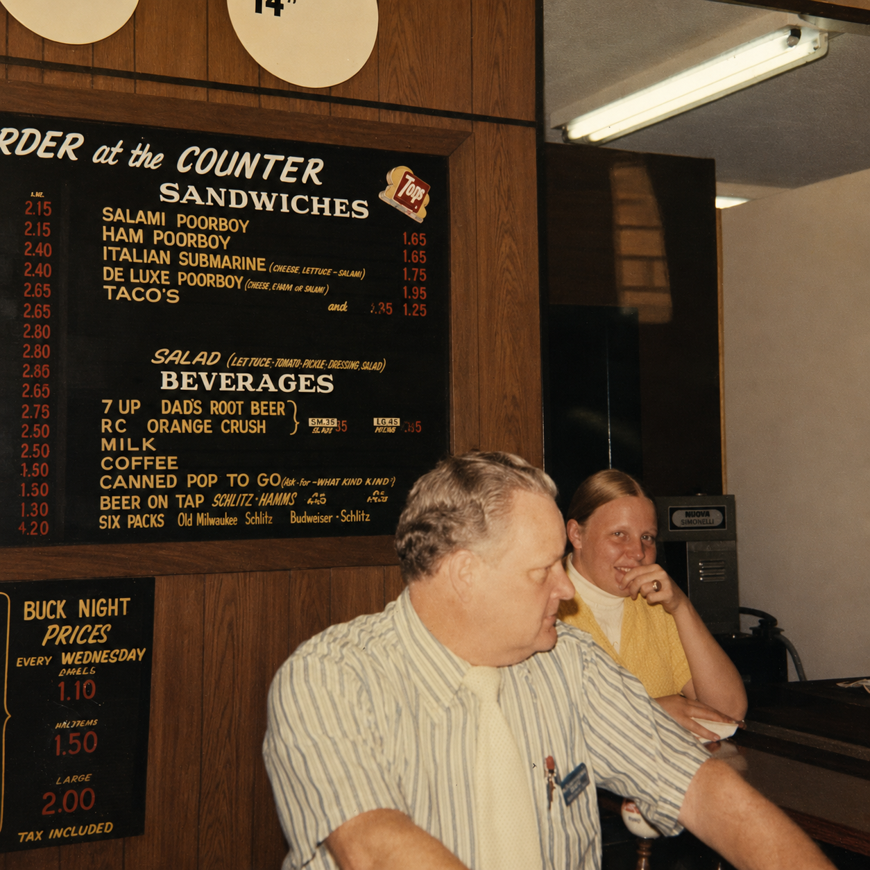 A blackboard menu with food and beverage options in a restaurant, with two employees behind the counter. One woman is smiling, and a man is looking down, appearing to work.