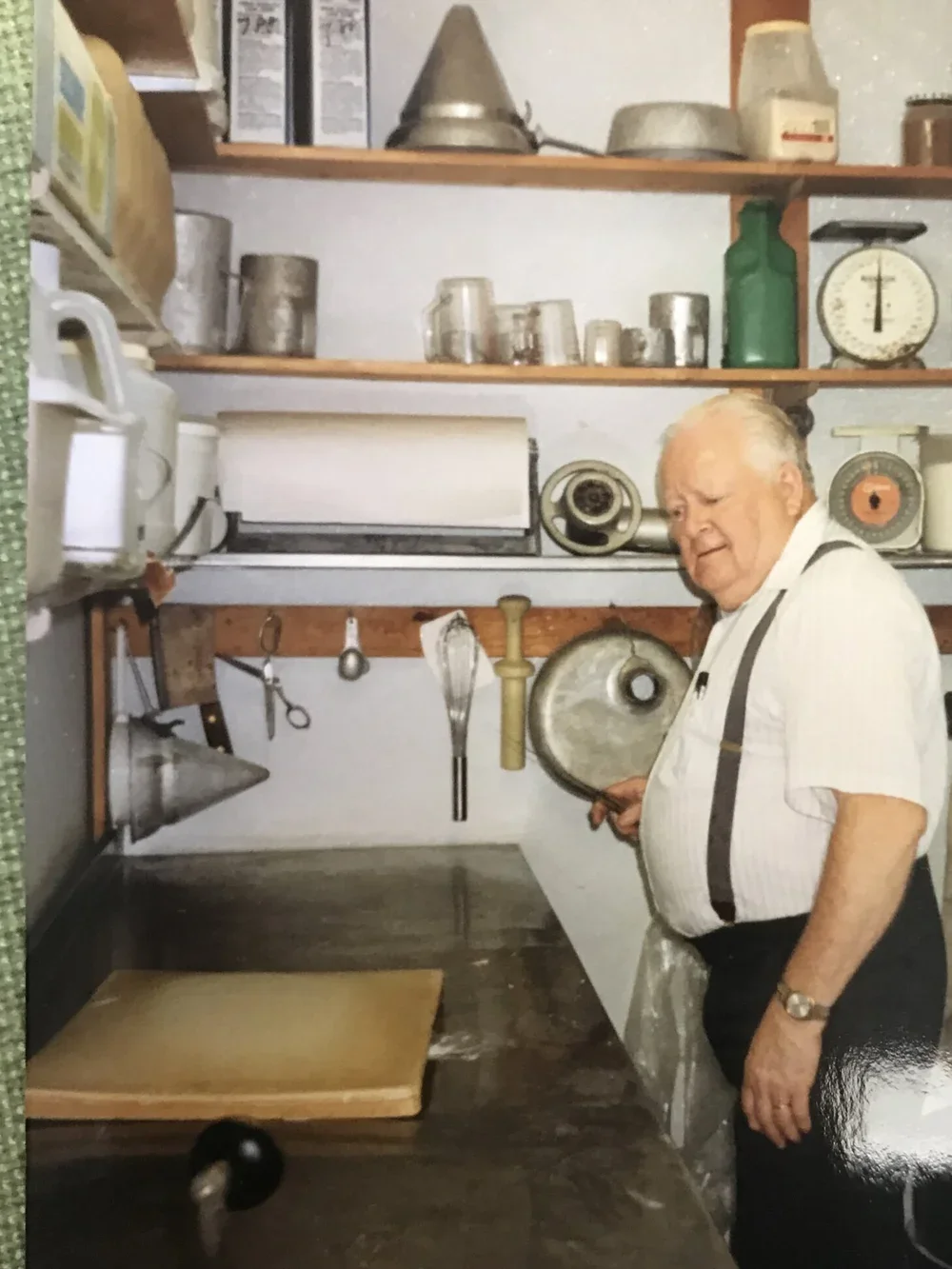An elderly man in a striped shirt and suspenders baking in a vintage kitchen, holding a pot lid in front of a metal counter with a rolling pin hanging on the wall and shelves with kitchen utensils and containers.