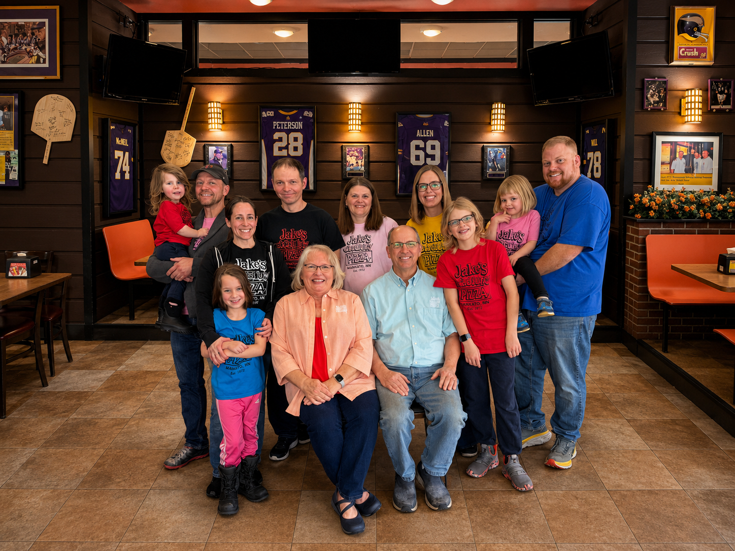 A group of fifteen people, including children, gathered in a restaurant decorated with sports jerseys and memorabilia, smiling for the photo.