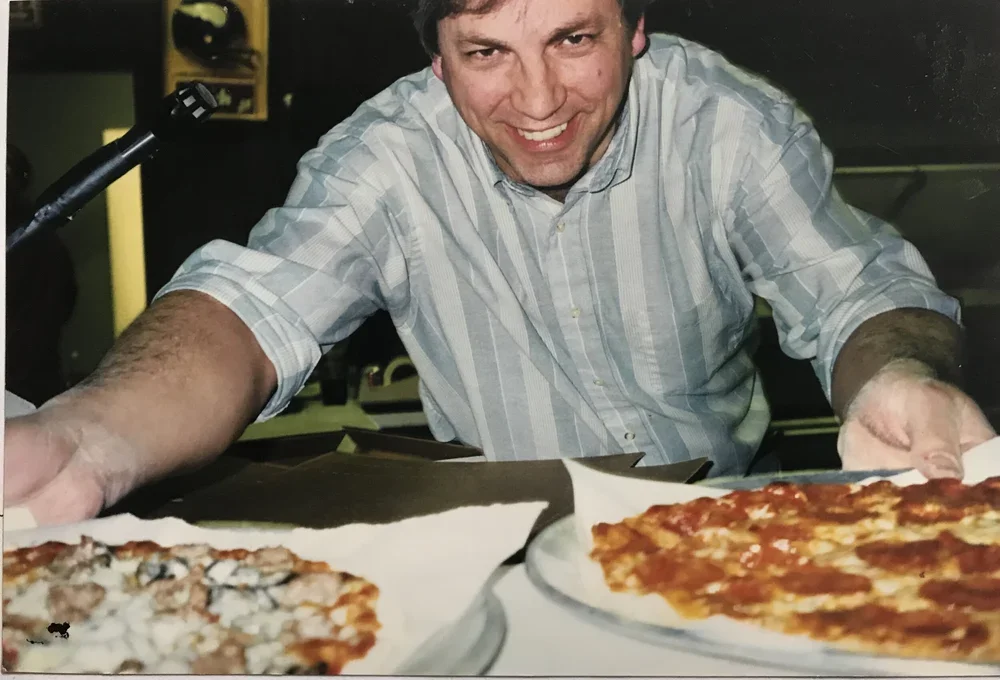 A man in a striped shirt smiling and reaching for pizza on a table.