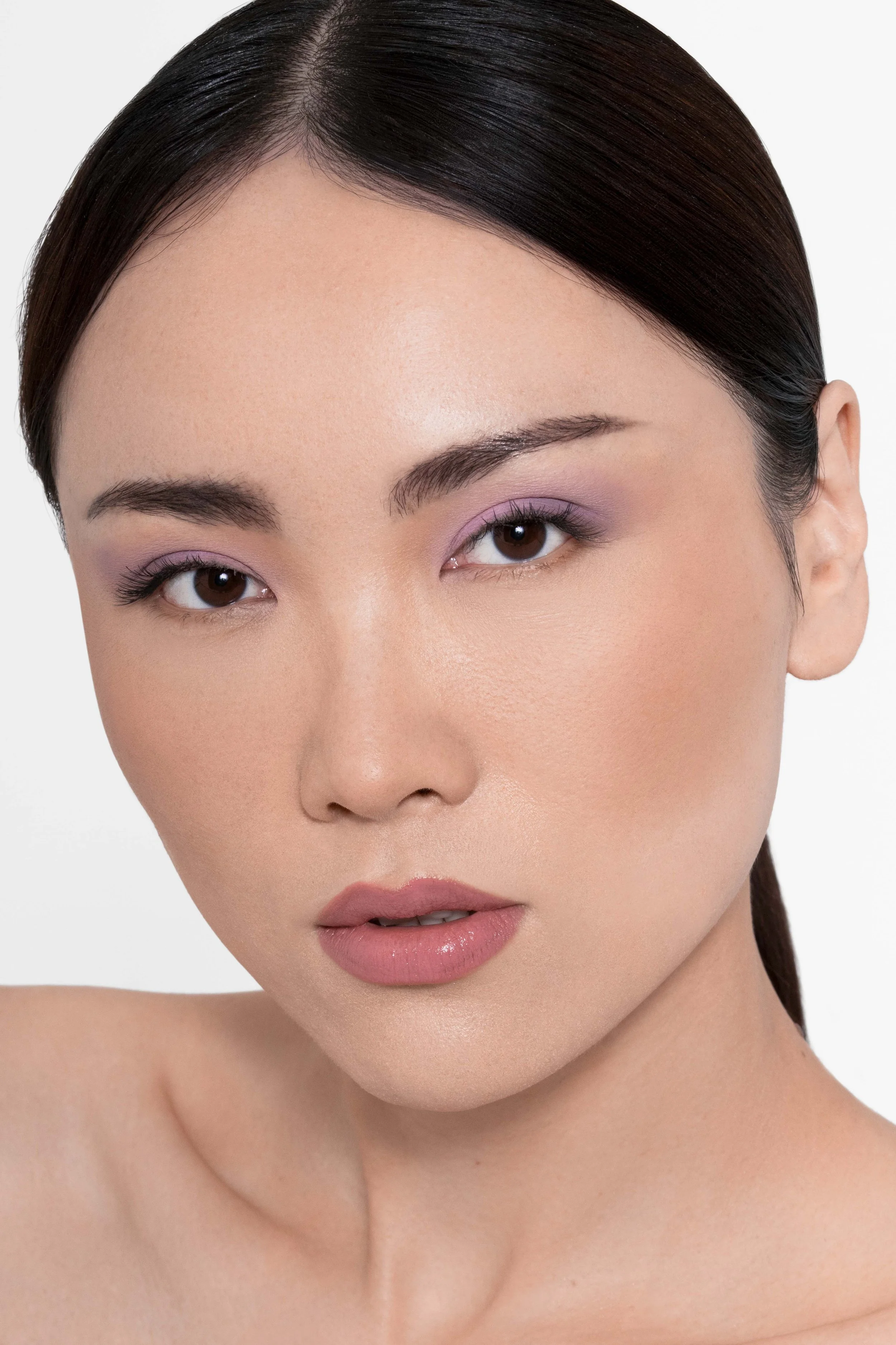 Beauty portrait of a model with sleek dark hair, luminous skin, soft pink and purple eye makeup, and natural nude lips. The clean white background and polished makeup create a fresh, sophisticated aesthetic, ideal for beauty campaigns, cosmetics adve
