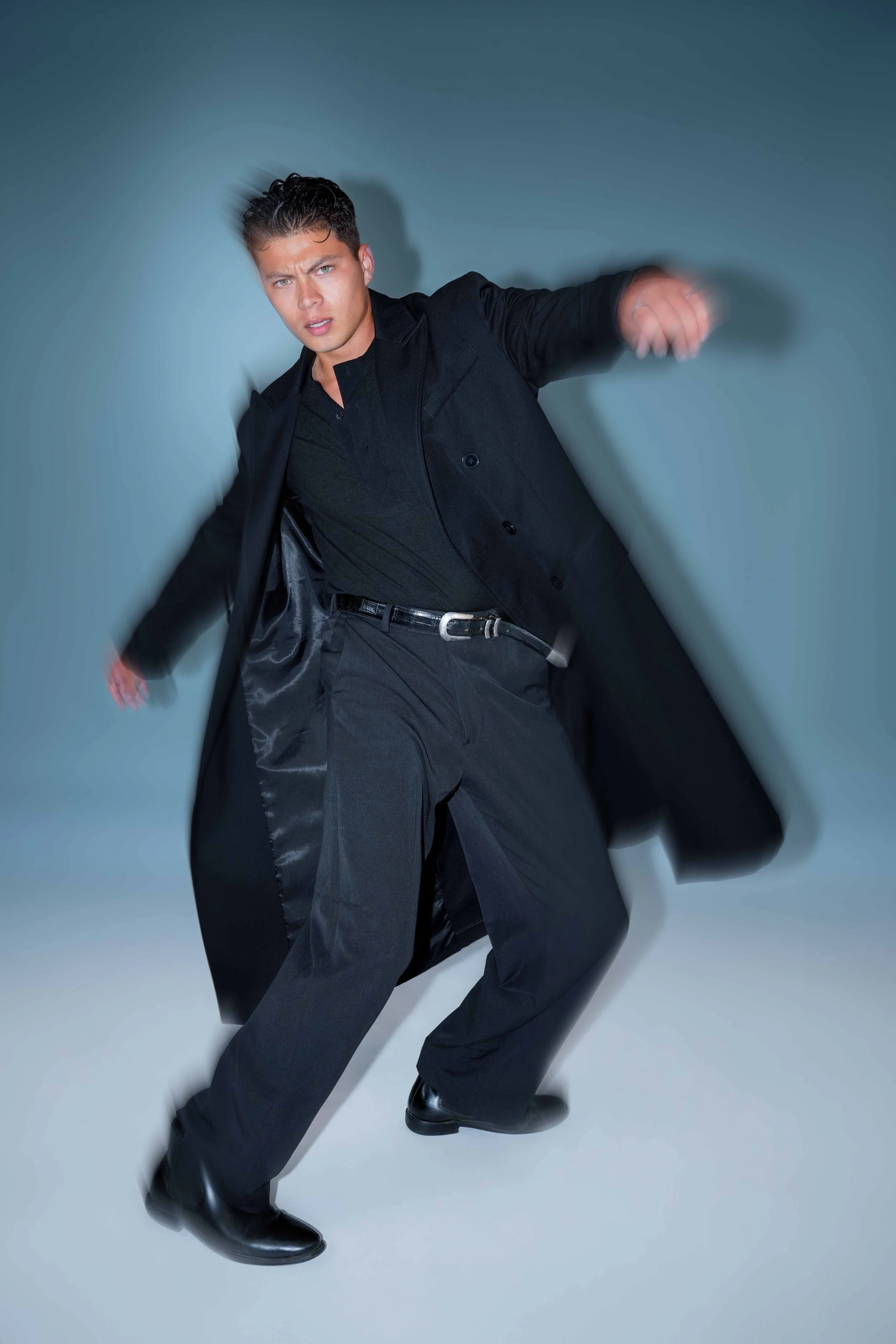 Male fashion model in motion wearing a black outfit and long coat during editorial photoshoot.