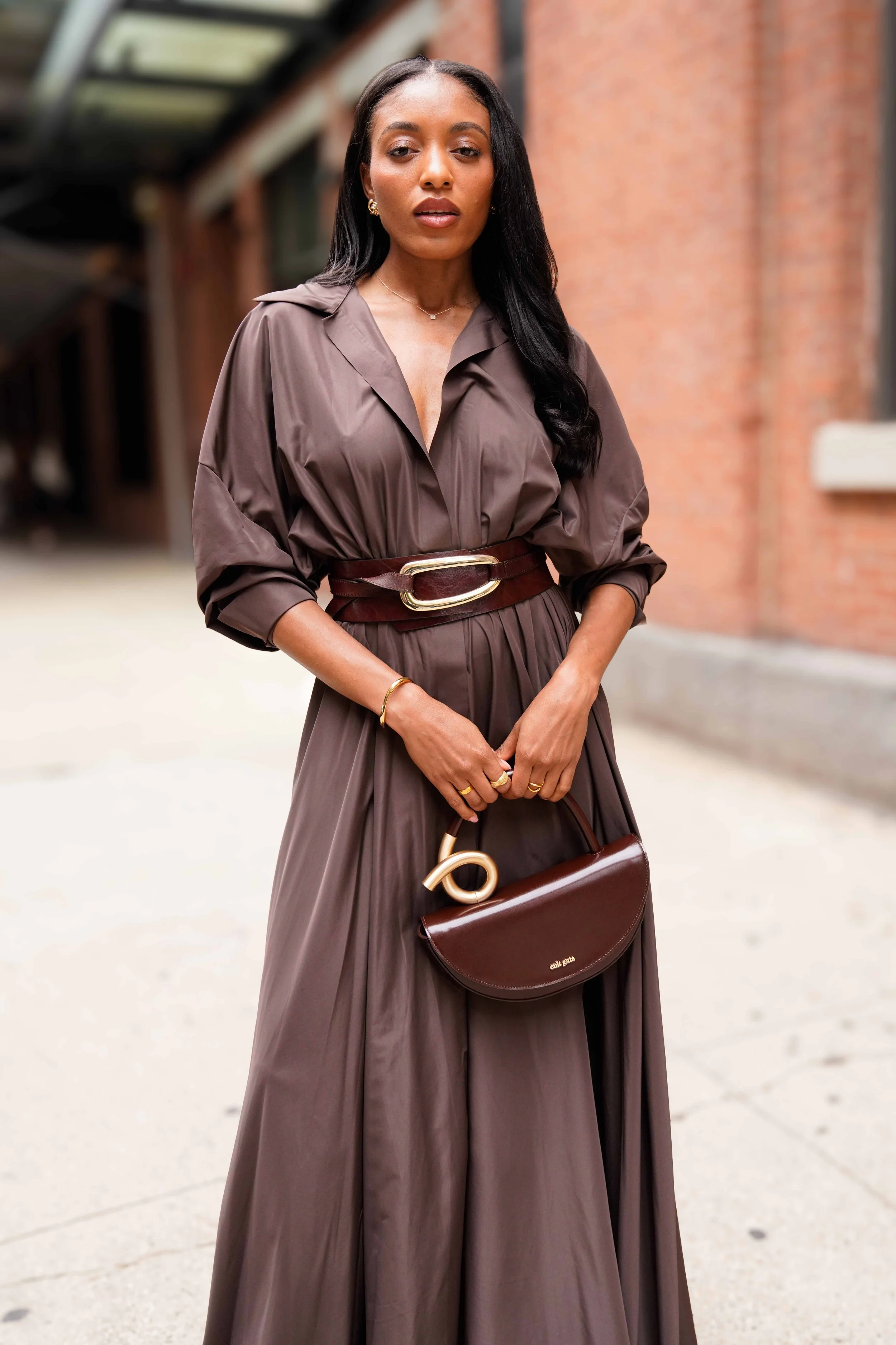 A woman in a long, brown dress and matching belt stands outdoors, holding a brown handbag, against a backdrop of brick buildings.