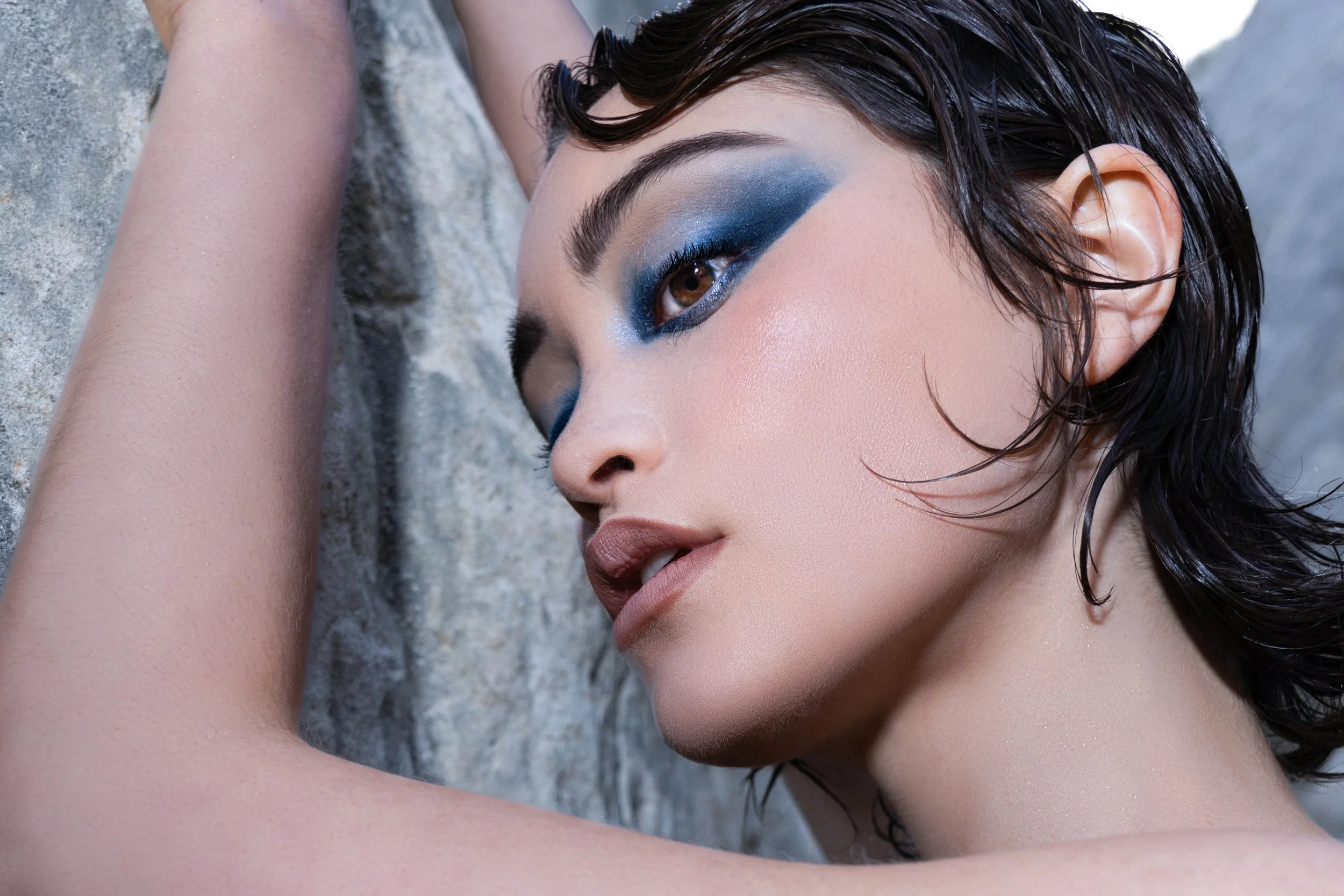 Close-up beauty portrait of a fashion model with dramatic blue editorial eye makeup and wet-look hair photographed against natural canyon rock textures. The image highlights flawless glowing skin, sharp facial structure, and intense eye contact, crea