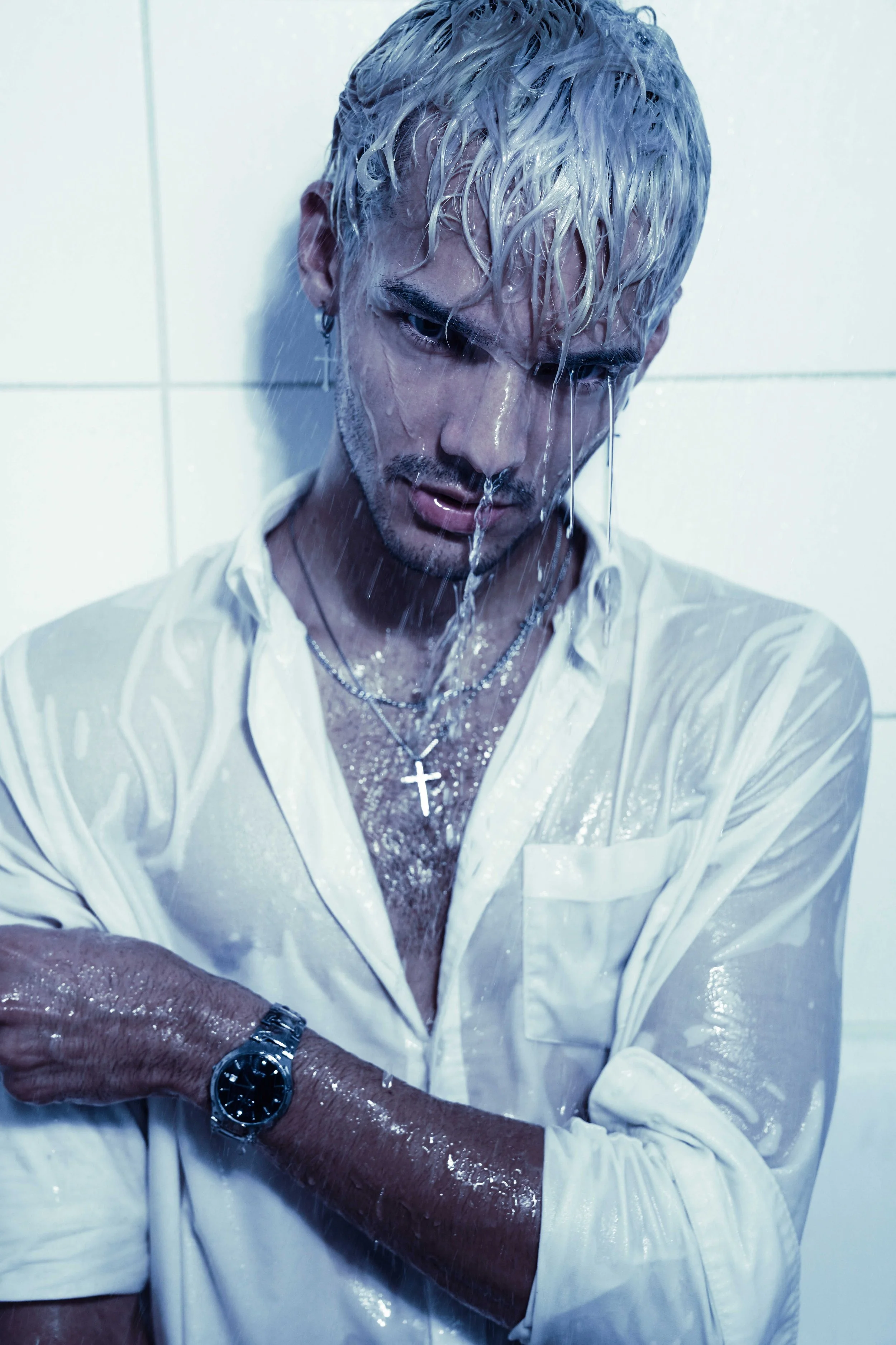 Blonde male model is posing in a bathtub, being wet, wearing a white button-down shirt and silver jewelry.