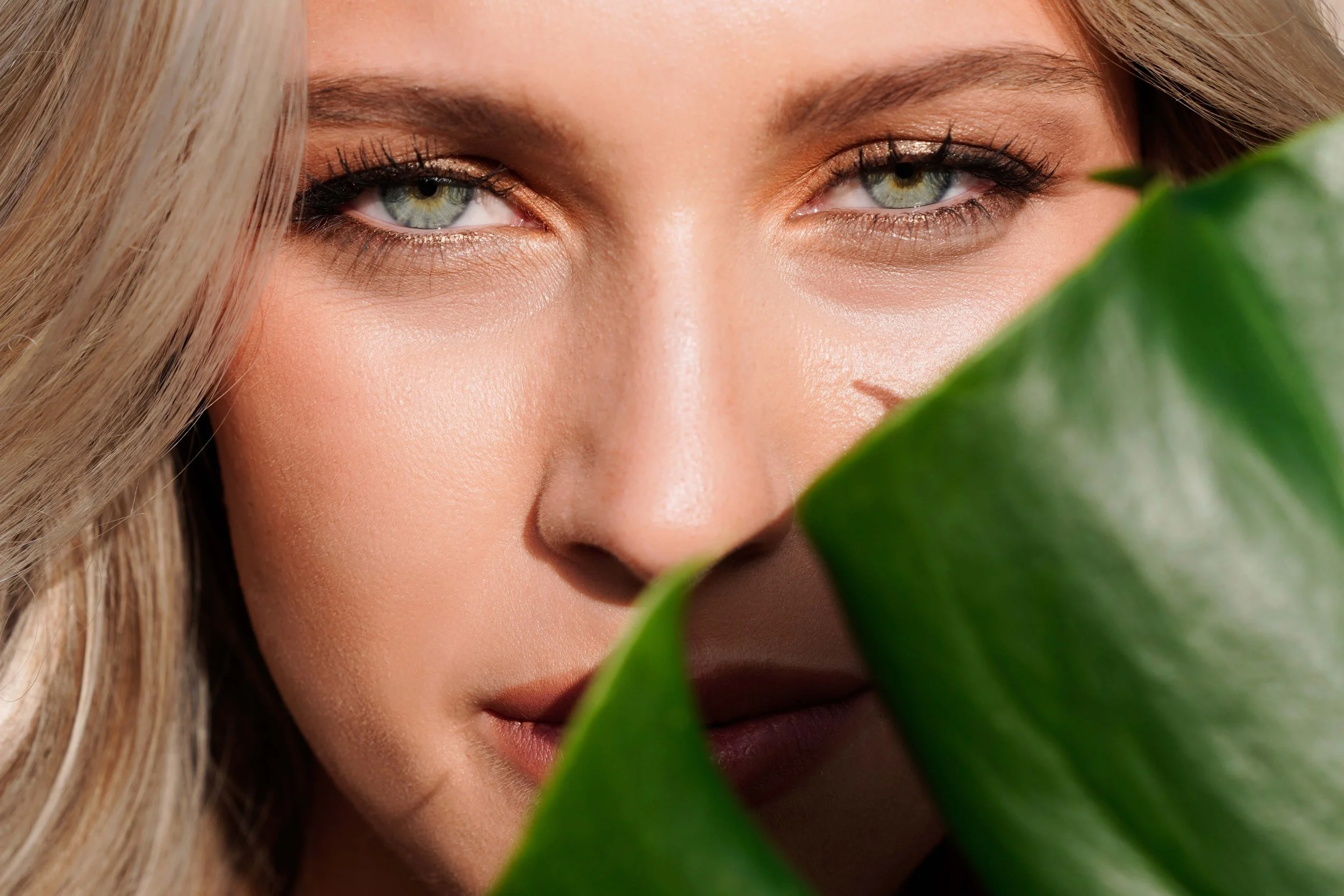 High-end close-up beauty photography of a blonde female model with radiant skin and piercing blue eyes, partially concealed by a green tropical leaf. This editorial studio portrait blends soft natural beauty, refined retouching, and bold composition 