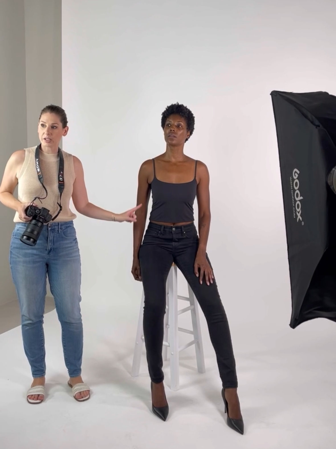 A woman is sitting on a stool in a photo studio, with a photographer on her left pointing and explaining. The woman is wearing a black spaghetti strap top, black jeans, and high heels. A large softbox light is set up to the right.
