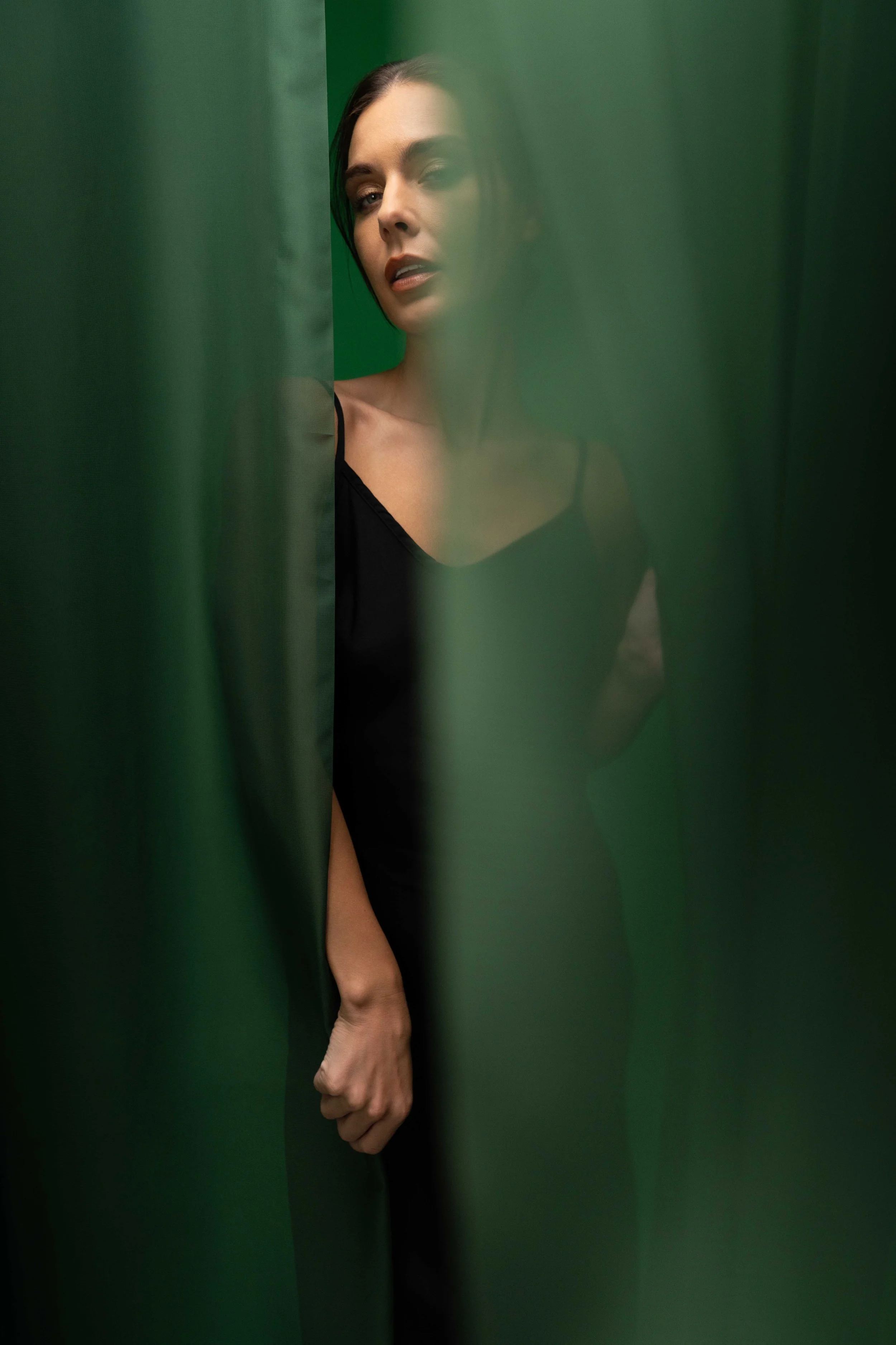 Moody editorial portrait of a fashion model framed between flowing emerald green fabric panels, creating a cinematic and mysterious atmosphere. The soft blurred foreground, elegant black dress, and subtle expression give the image a modern luxury fas