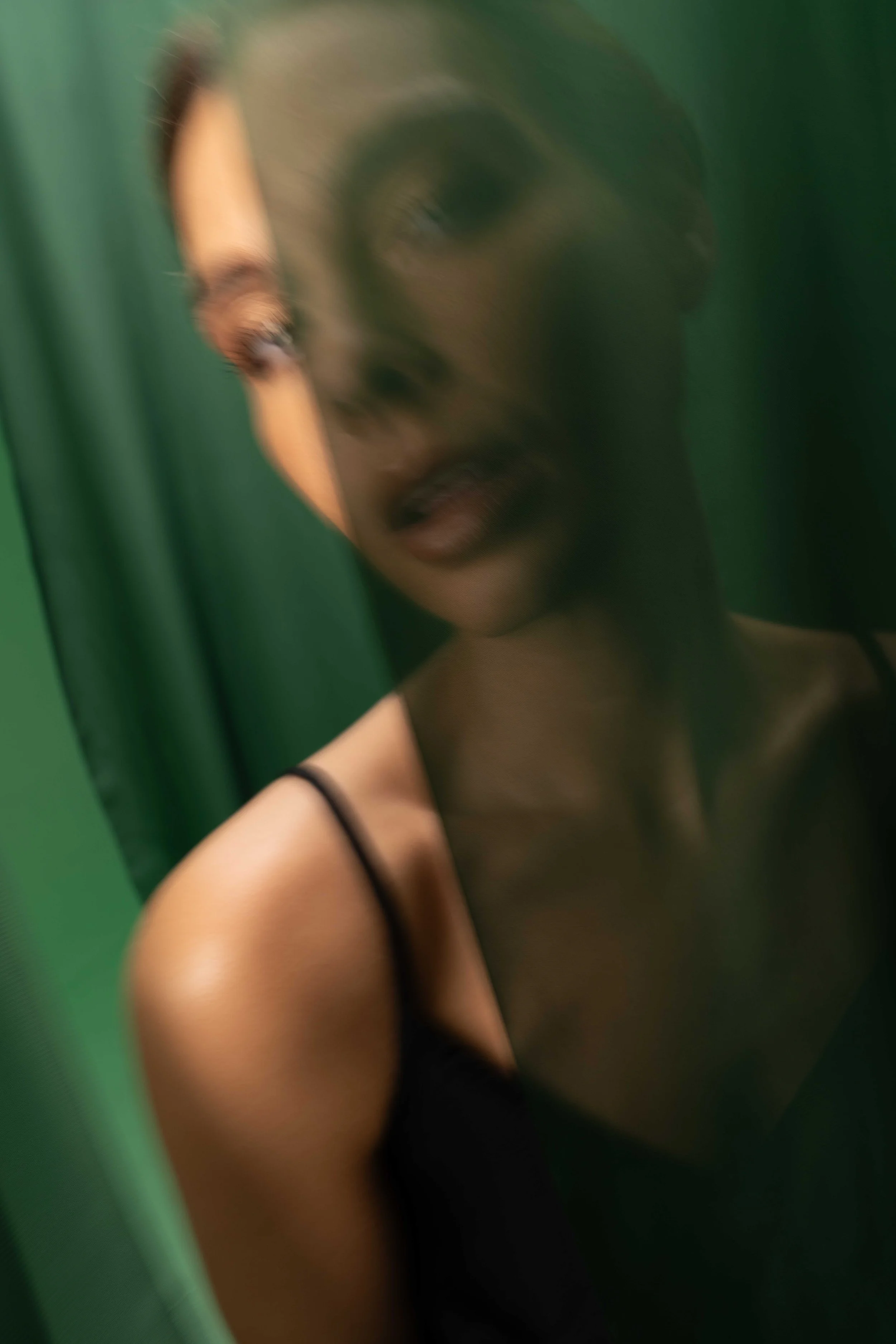 Moody editorial portrait of a fashion model framed between flowing emerald green fabric panels, creating a cinematic and mysterious atmosphere. The soft blurred foreground, elegant black dress, and subtle expression give the image a modern luxury fas