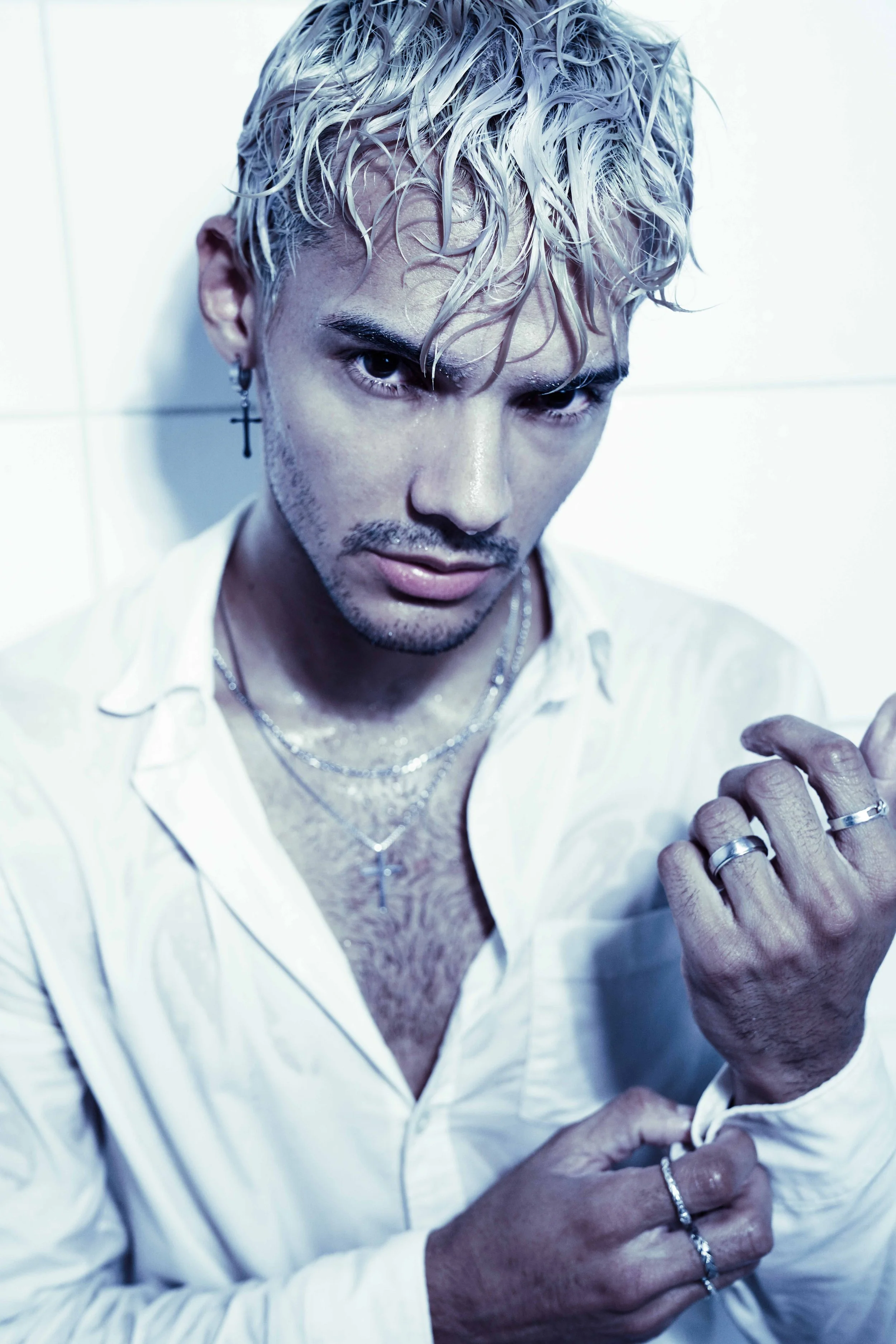 Blonde male model is posing in a bathtub, wearing a white button-down shirt and silver jewelry.