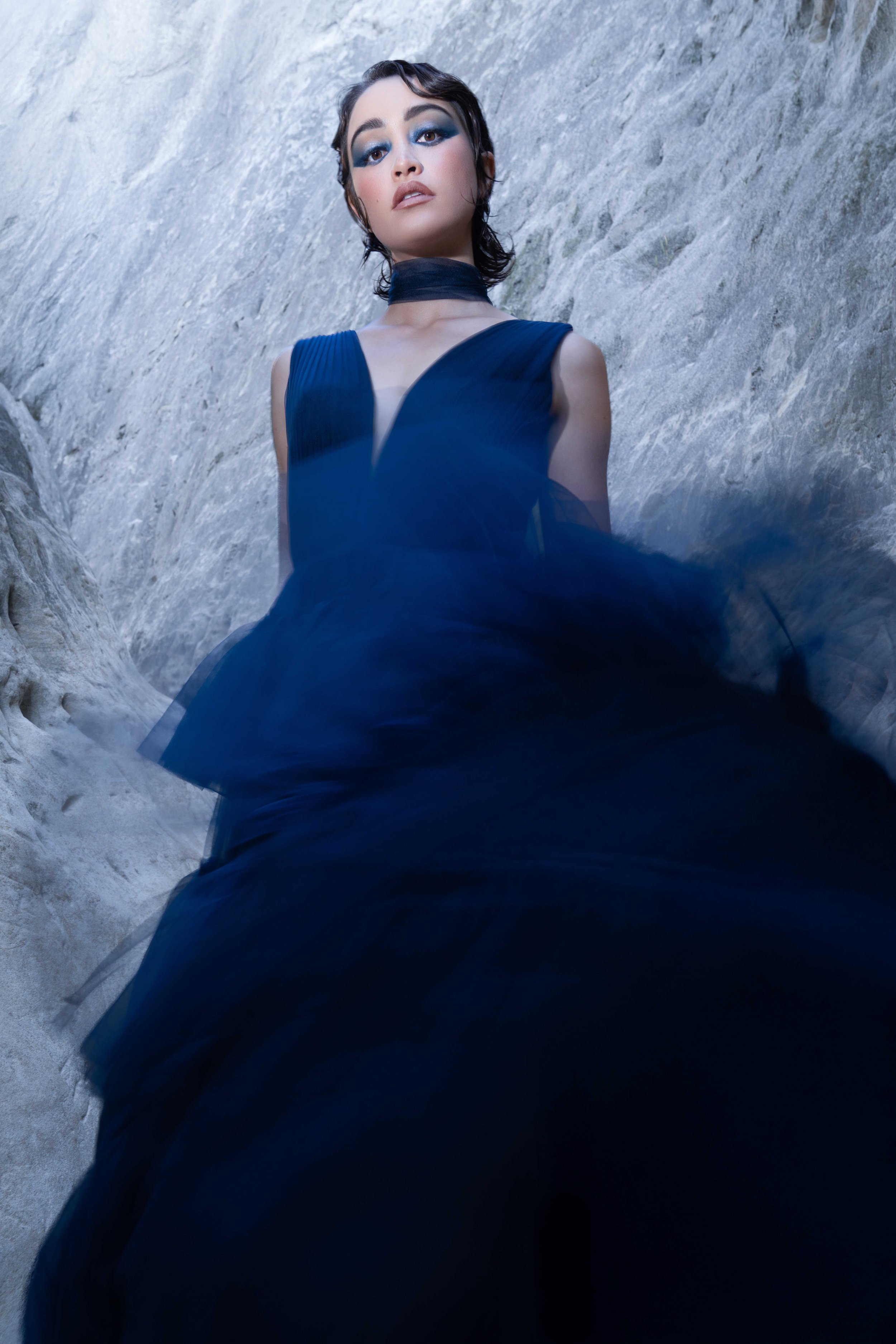 A fashion model wearing a flowing dark navy couture gown standing between dramatic canyon rock formations during a cinematic editorial fashion photoshoot. The low camera angle, movement in the sheer fabric, and confident expression create a powerful 