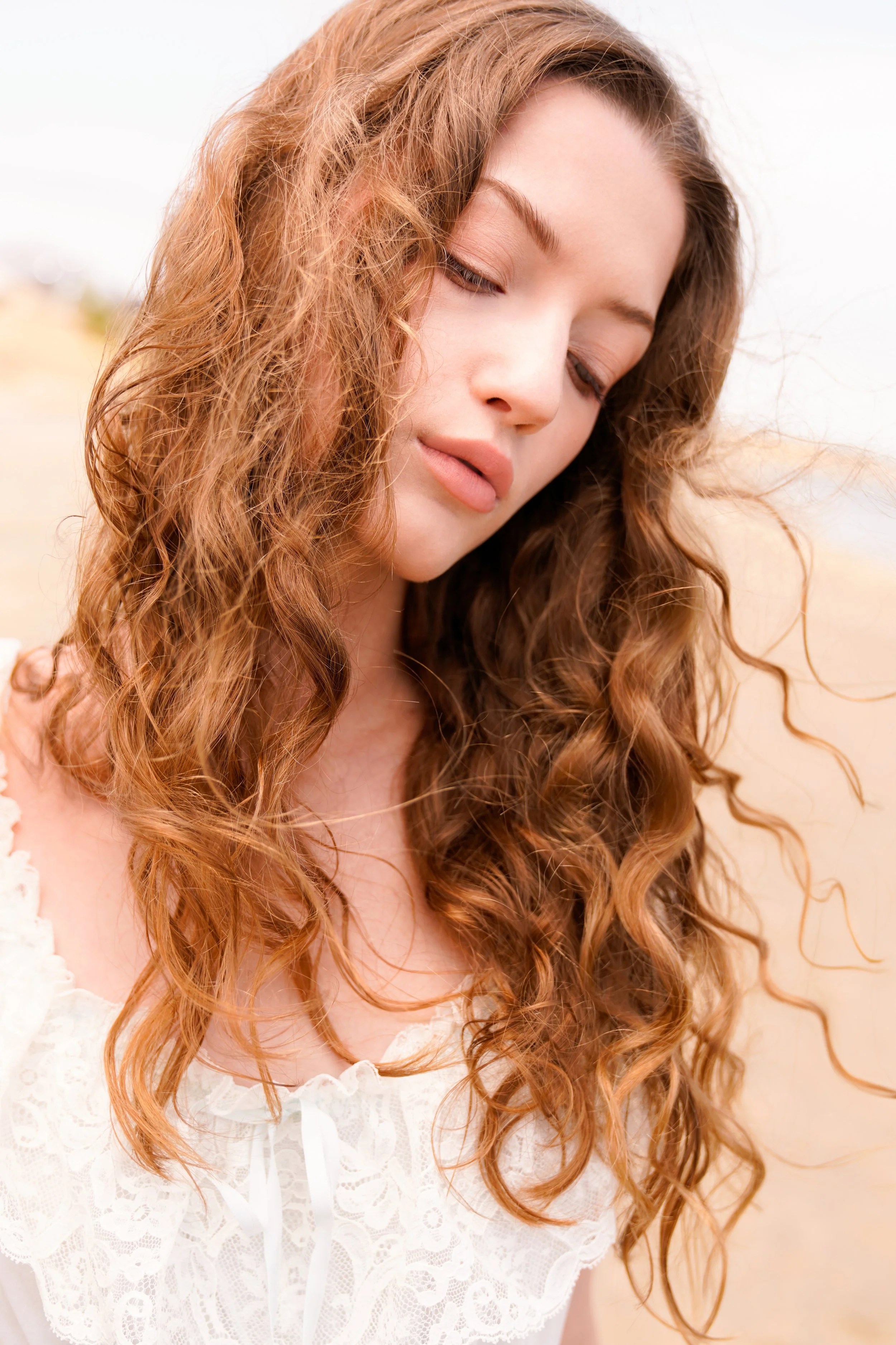 Close-up portrait of a young woman with natural makeup and long, curly auburn hair, posing with her arms raised under soft daylight. Captured by fashion and beauty photographer Sylvia Binder, this ethereal outdoor shot highlights effortless elegance,