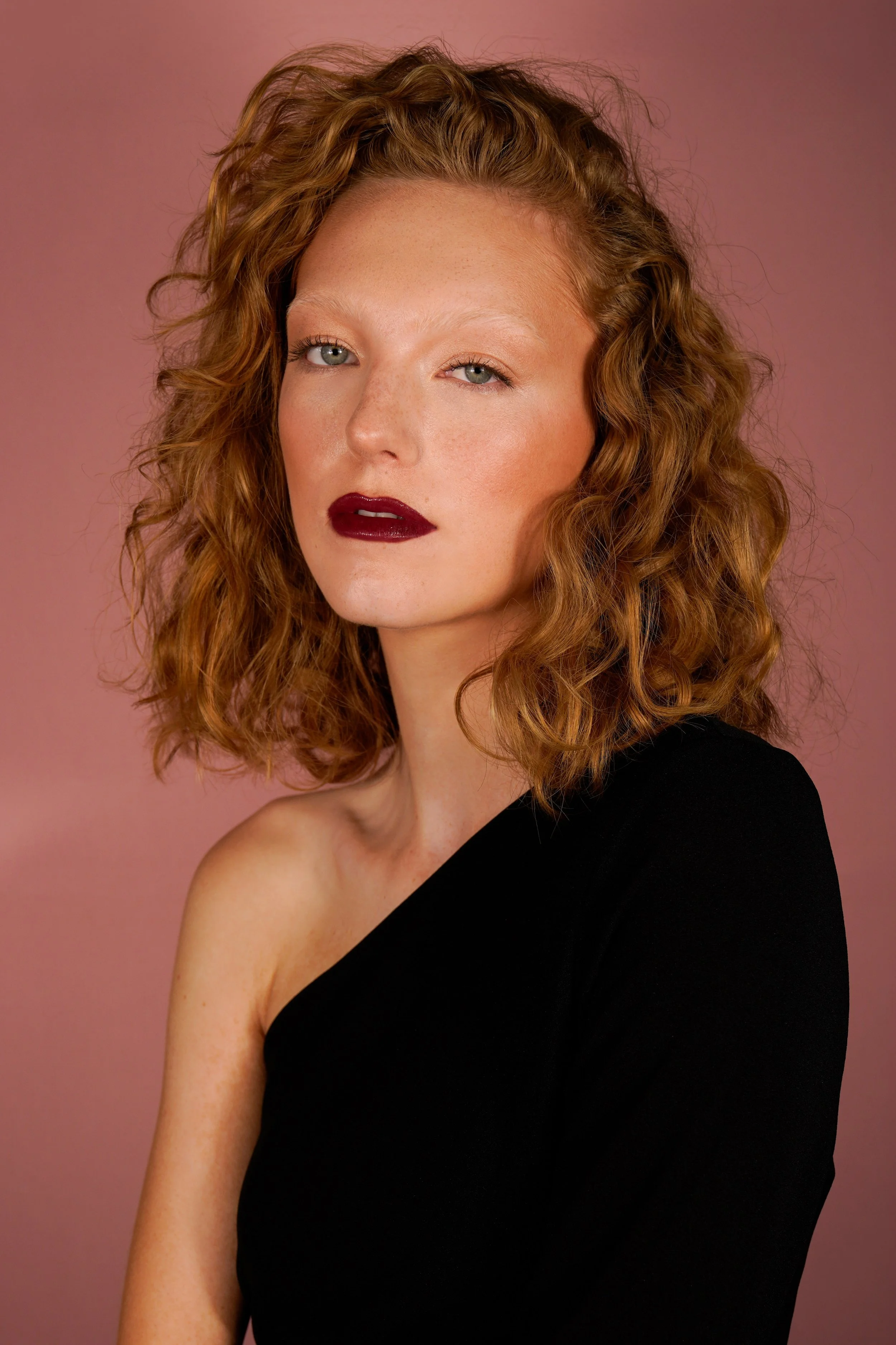 High-fashion beauty portrait of a red-haired model wearing a bold dark red lip, flawless glowing skin, and a one-shoulder black outfit. Set against a muted pink backdrop and captured by fashion and beauty photographer Sylvia Binder, this editorial im