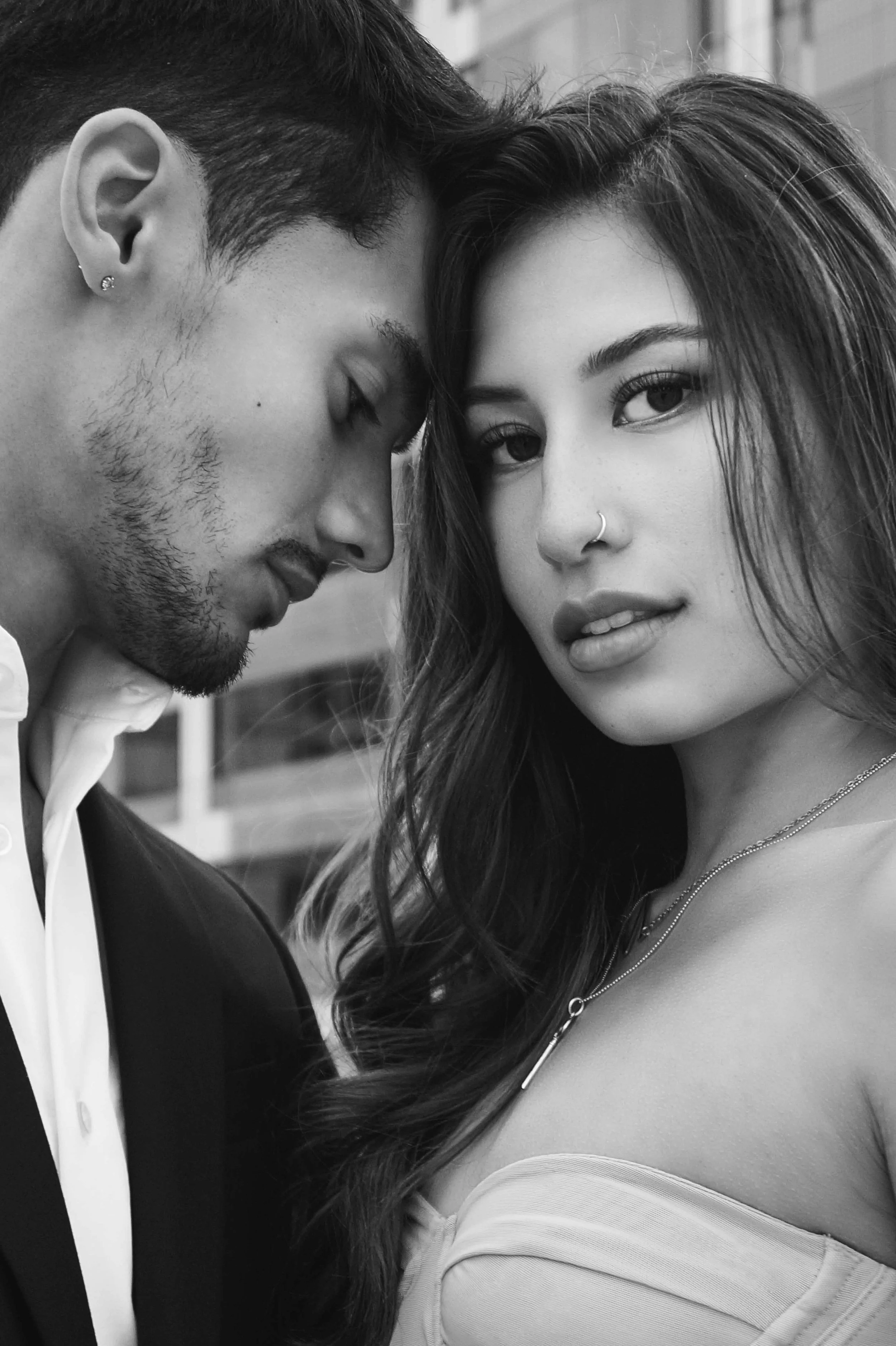 A black and white close-up photo of a young man with closed eyes leaning his forehead against a young woman with long wavy hair, both facing the camera. The woman has light skin, dark eyes, a nose ring, and wears a strapless top and necklace.