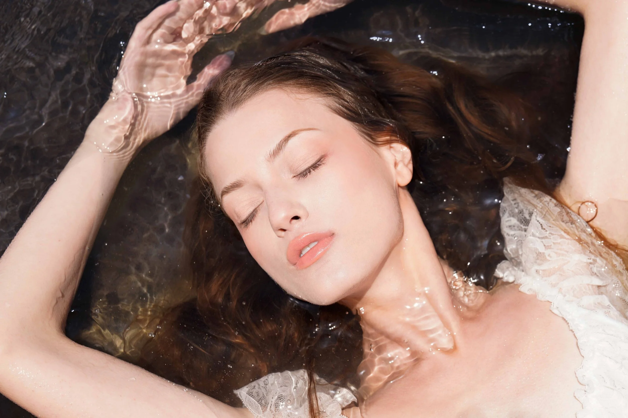 Close-up beauty portrait of a young woman with glowing skin and natural makeup, floating in water with her eyes closed. Captured by fashion and beauty photographer Sylvia Binder, this serene and ethereal water shoot highlights soft textures, dreamy l