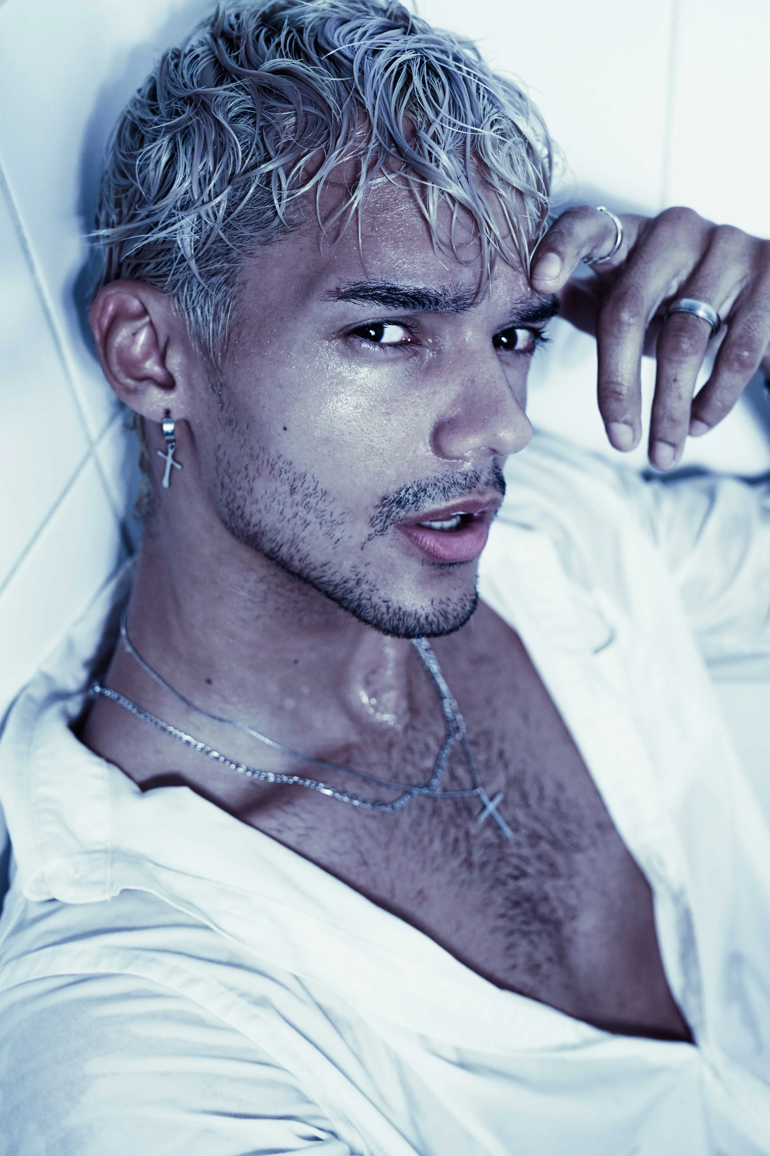 Blonde male model is posing in a bathtub, being wet, wearing a white button-down shirt and silver jewelry.