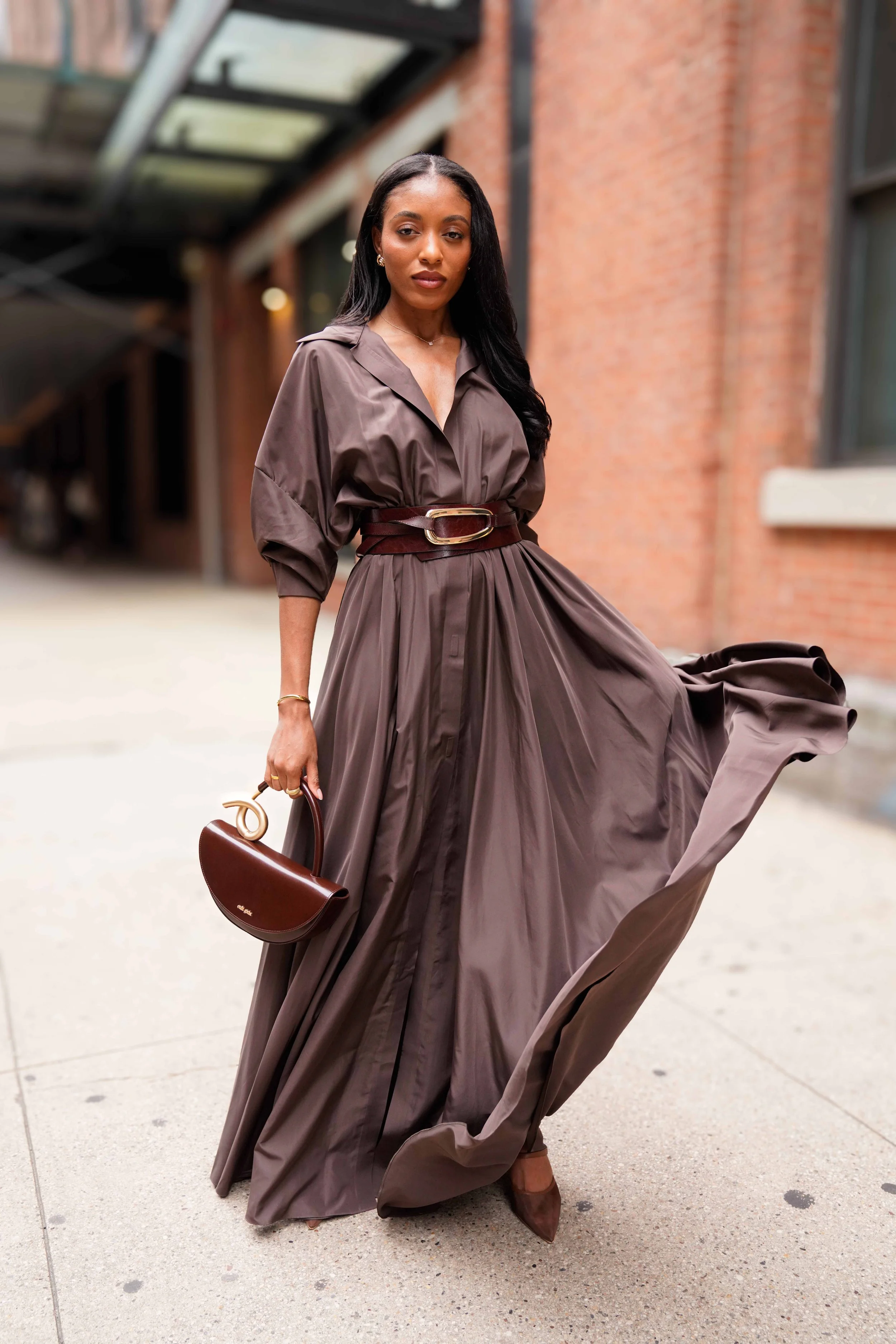 Woman wearing a brown long dress with a belt, holding a brown handbag, standing on a city sidewalk near a brick building.