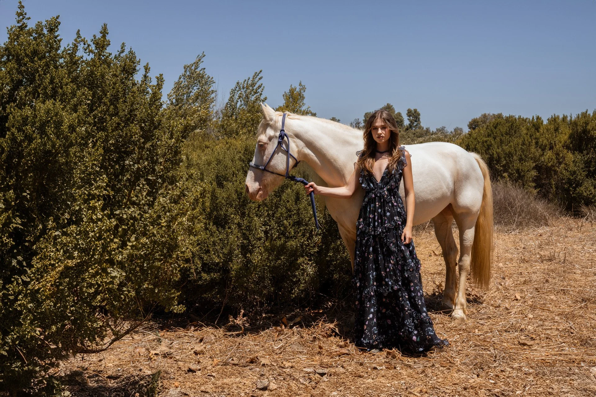 Editorial fashion photography of a model in a blue floral dress posing with a white horse in a rustic outdoor landscape in California.