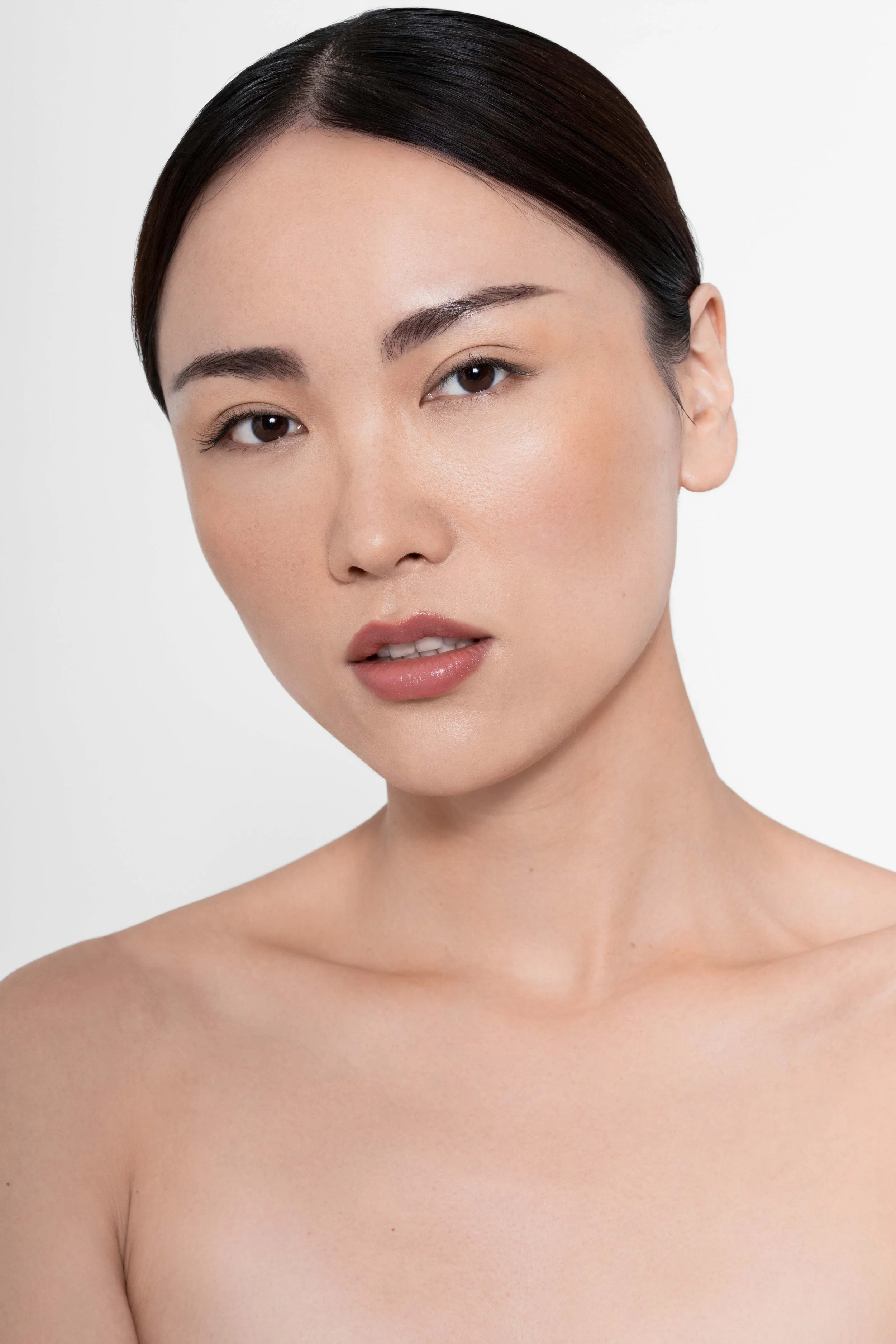 Beauty portrait of a model with sleek dark hair, luminous skin, natural makeup, and natural nude lips. The clean white background and polished makeup create a fresh, sophisticated aesthetic, ideal for beauty campaigns, cosmetics advertising, skincare