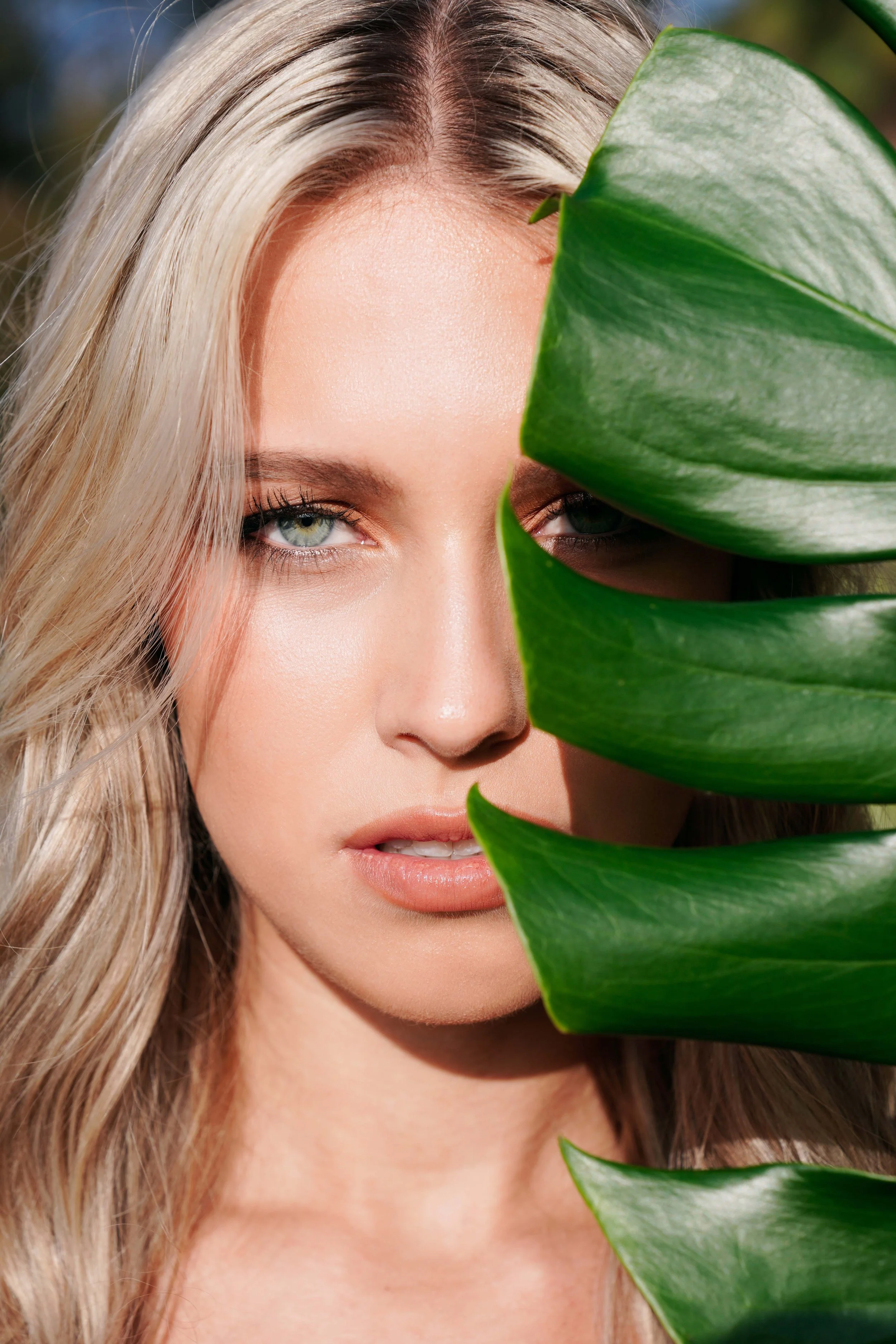 Professional beauty portrait of a blonde model partially framed by tropical green leaves, showcasing luminous skin, minimal natural makeup, and striking blue eyes. This editorial beauty photography combines clean studio lighting with organic elements