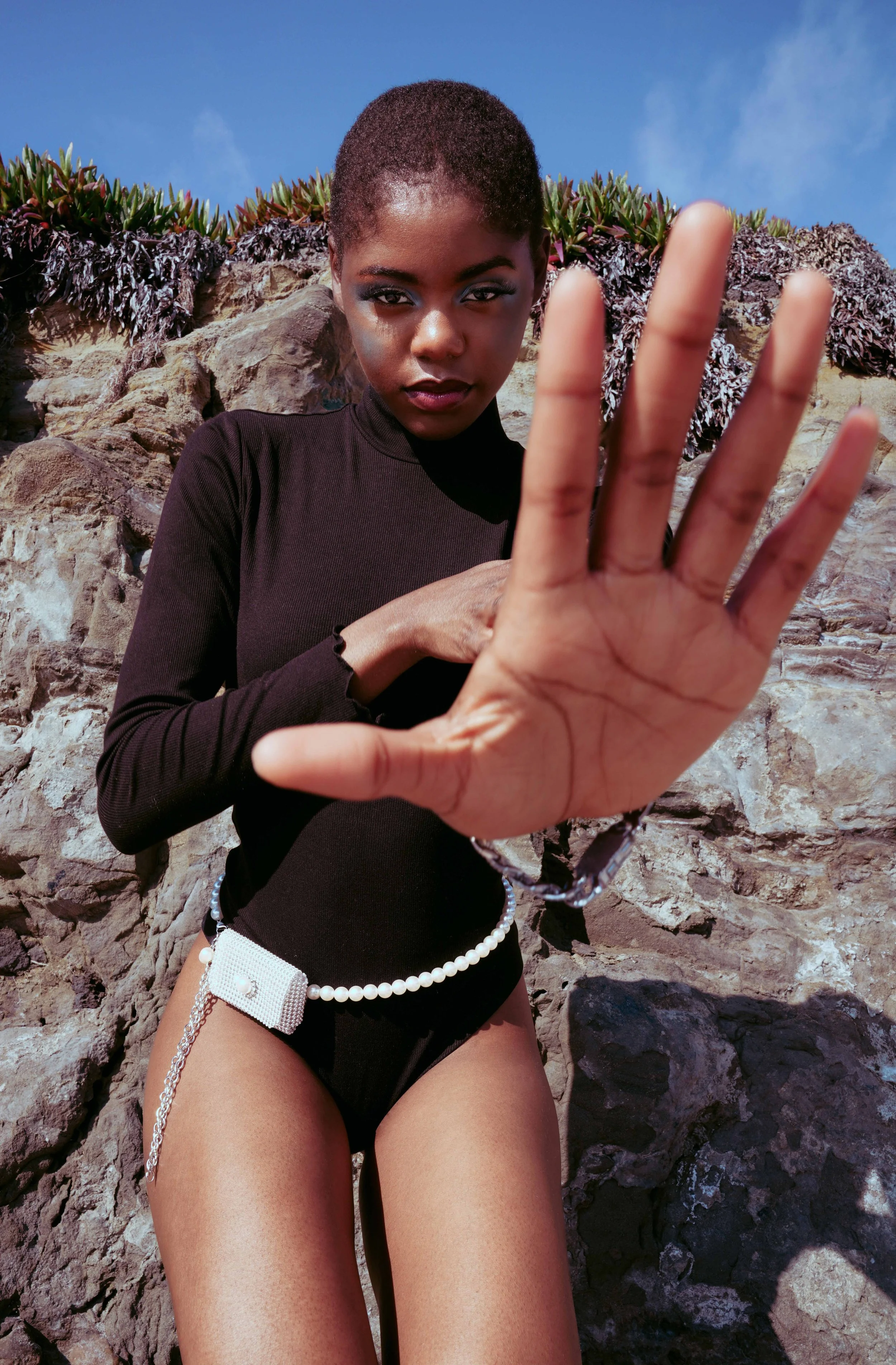 Black female model is posing in front of a brown rock, wearing a black body with a pearls purse belt.