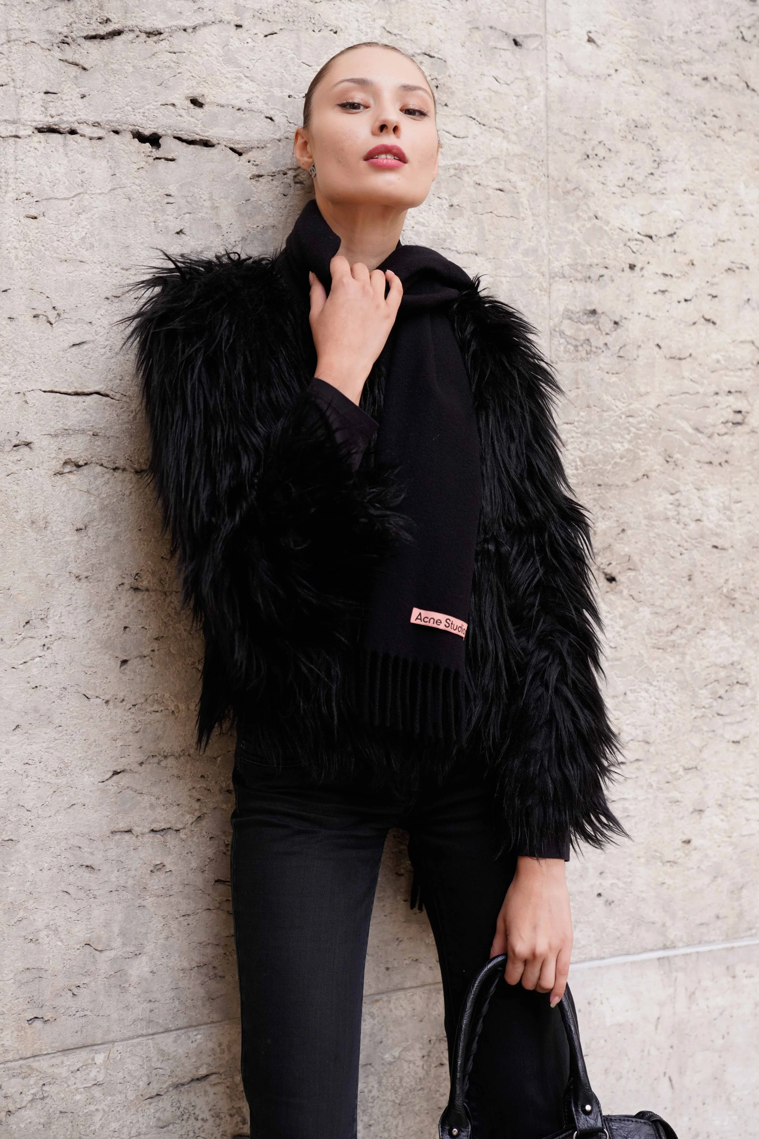 Woman wearing black faux fur jacket, black pants, and holding a black handbag against a beige textured wall.