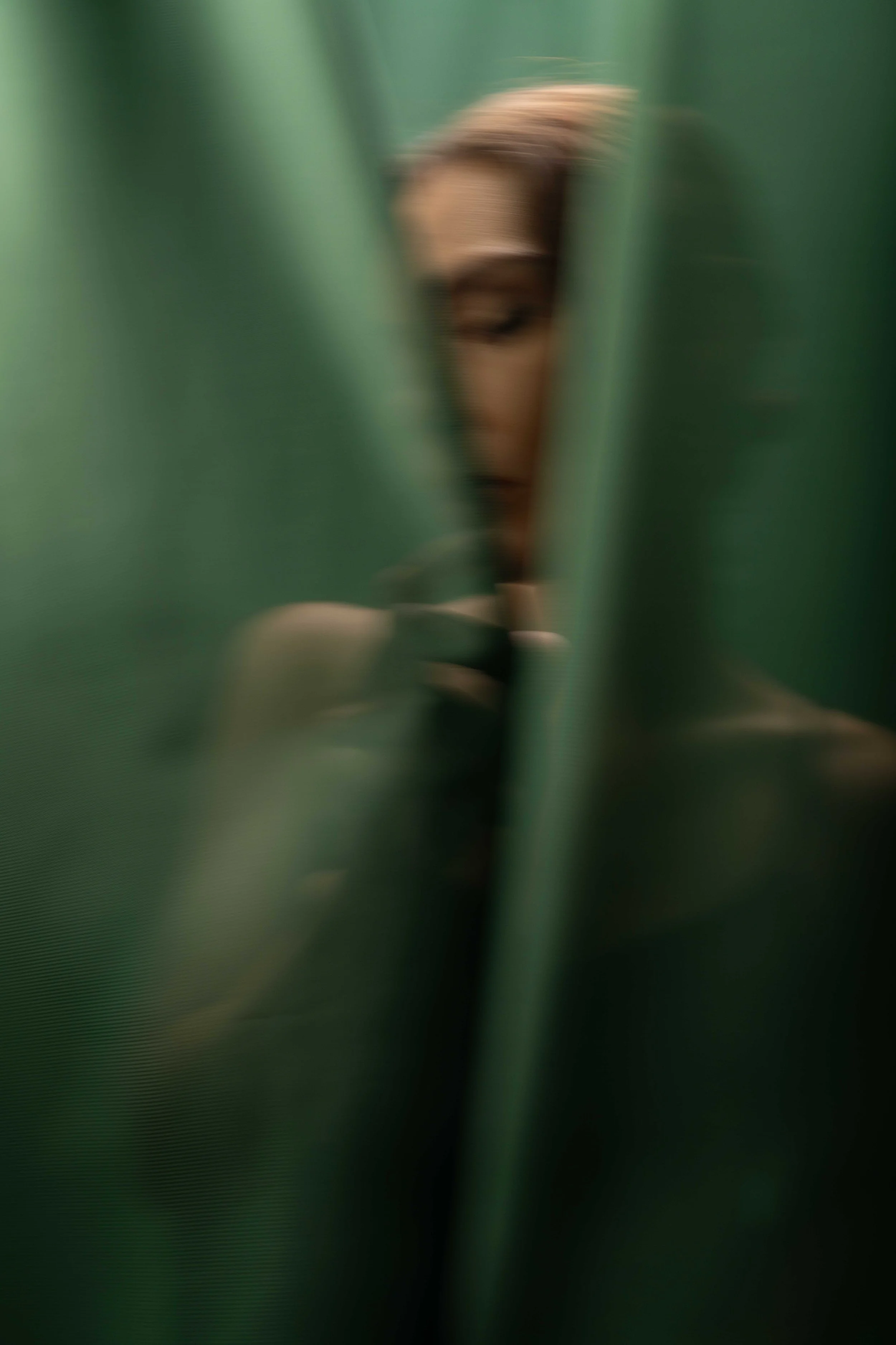 Moody editorial portrait of a fashion model framed between flowing emerald green fabric panels, creating a cinematic and mysterious atmosphere. The soft blurred foreground, elegant black dress, and subtle expression give the image a modern luxury fas