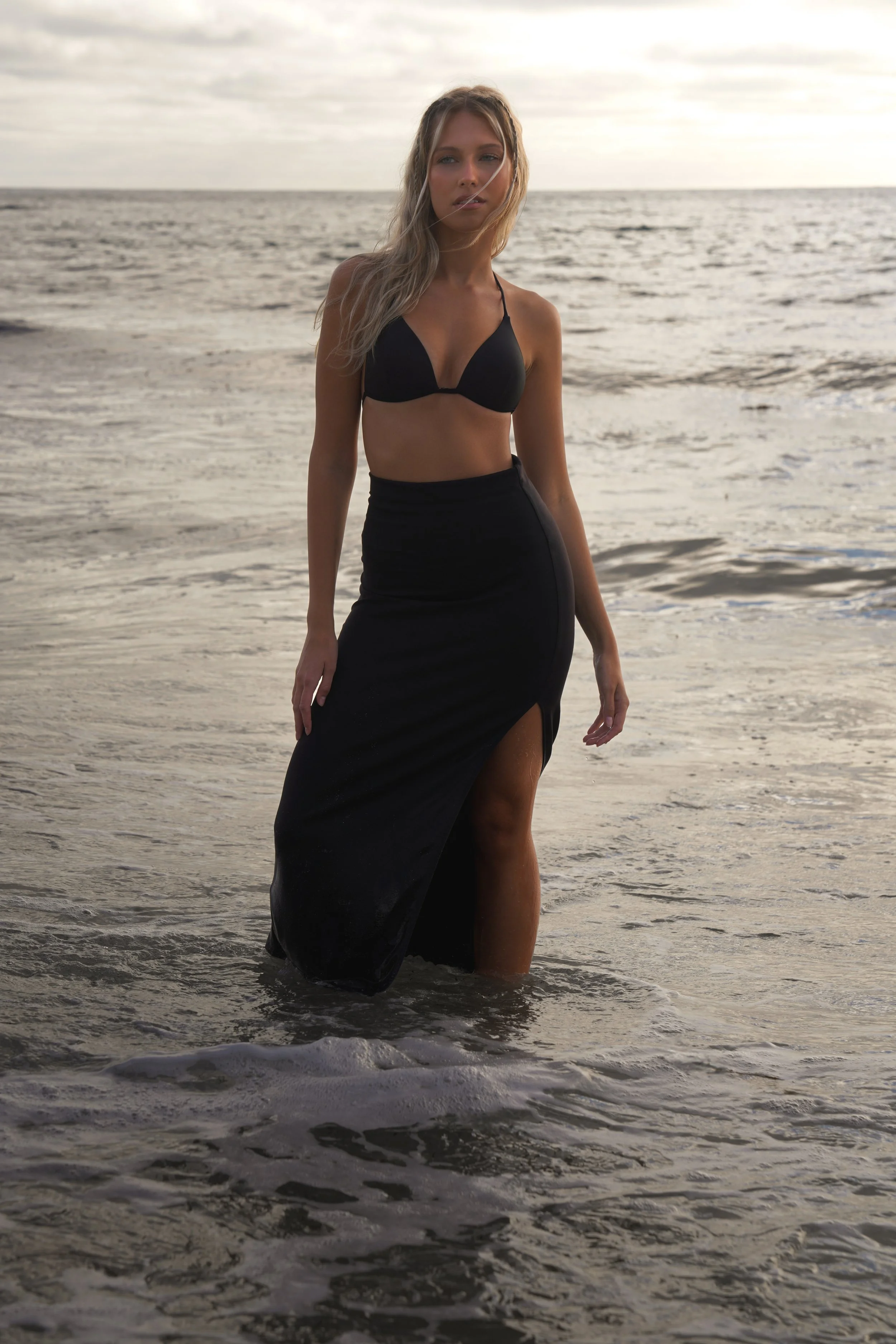Editorial fashion photoshoot at the beach. Blonde female model is posing in the water at sunset in a black bikini skirt set.