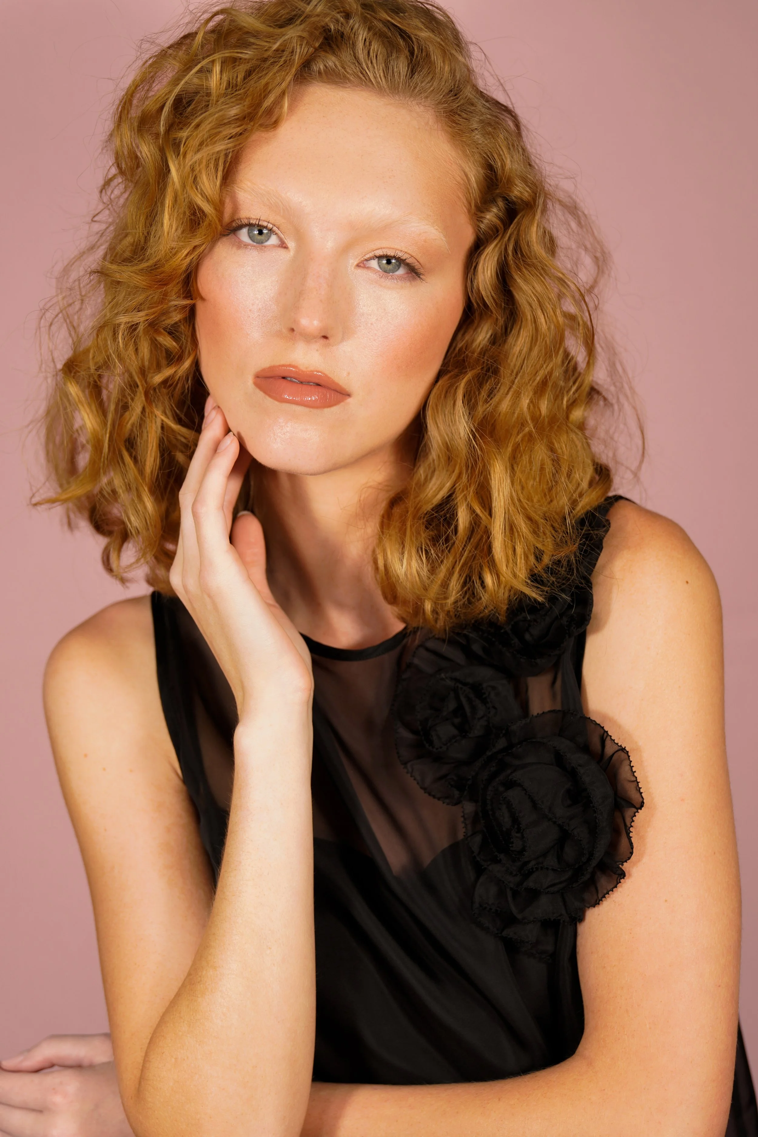 Studio beauty portrait of a red-haired model with glowing skin, soft glam makeup, and glossy nude lips, set against a muted pink background. Styled in a black sheer dress with floral appliqué, the image was captured by fashion and beauty photographer