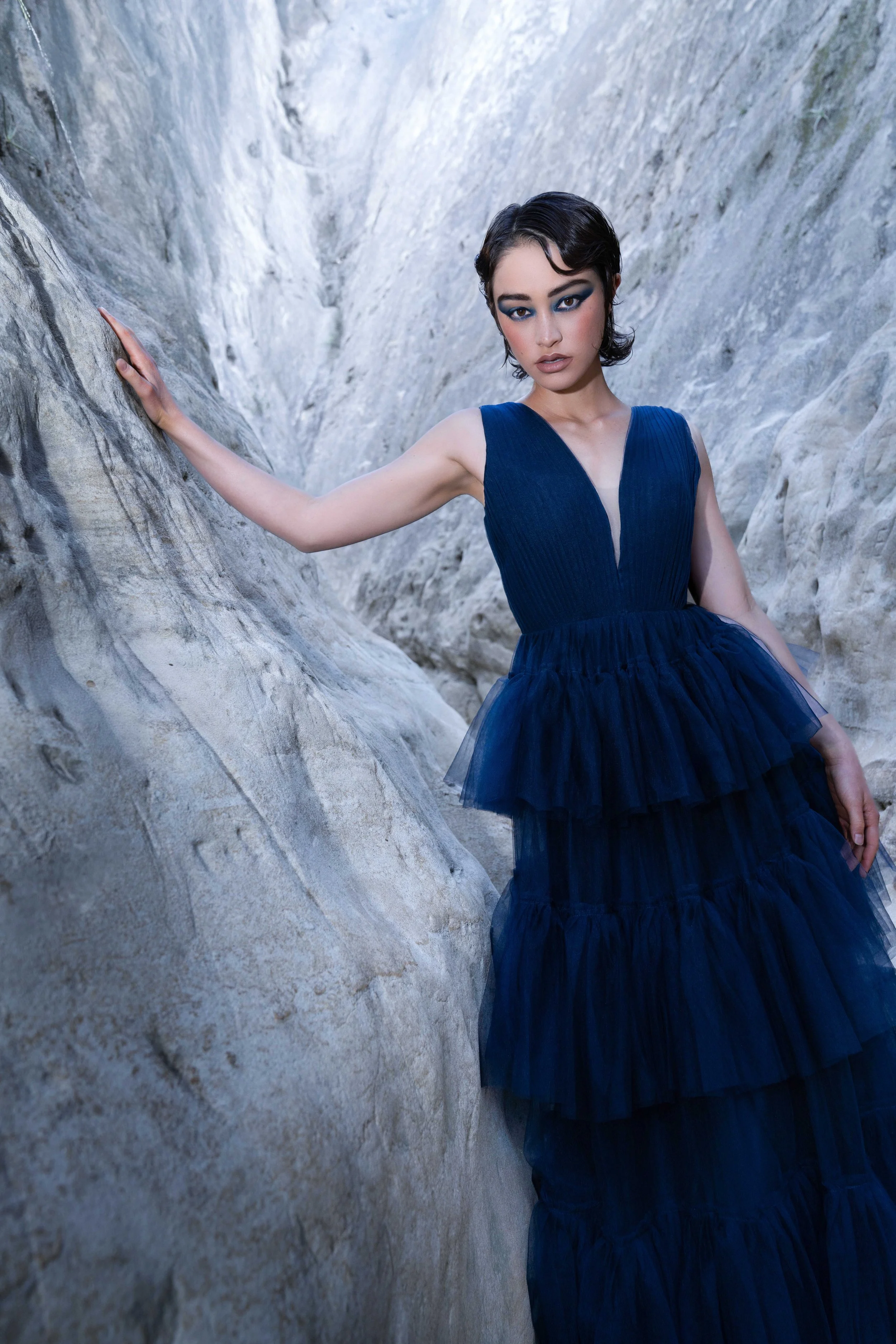 A fashion model wearing a flowing dark navy couture gown standing between dramatic canyon rock formations during a cinematic editorial fashion photoshoot. The low camera angle, movement in the sheer fabric, and confident expression create a powerful 