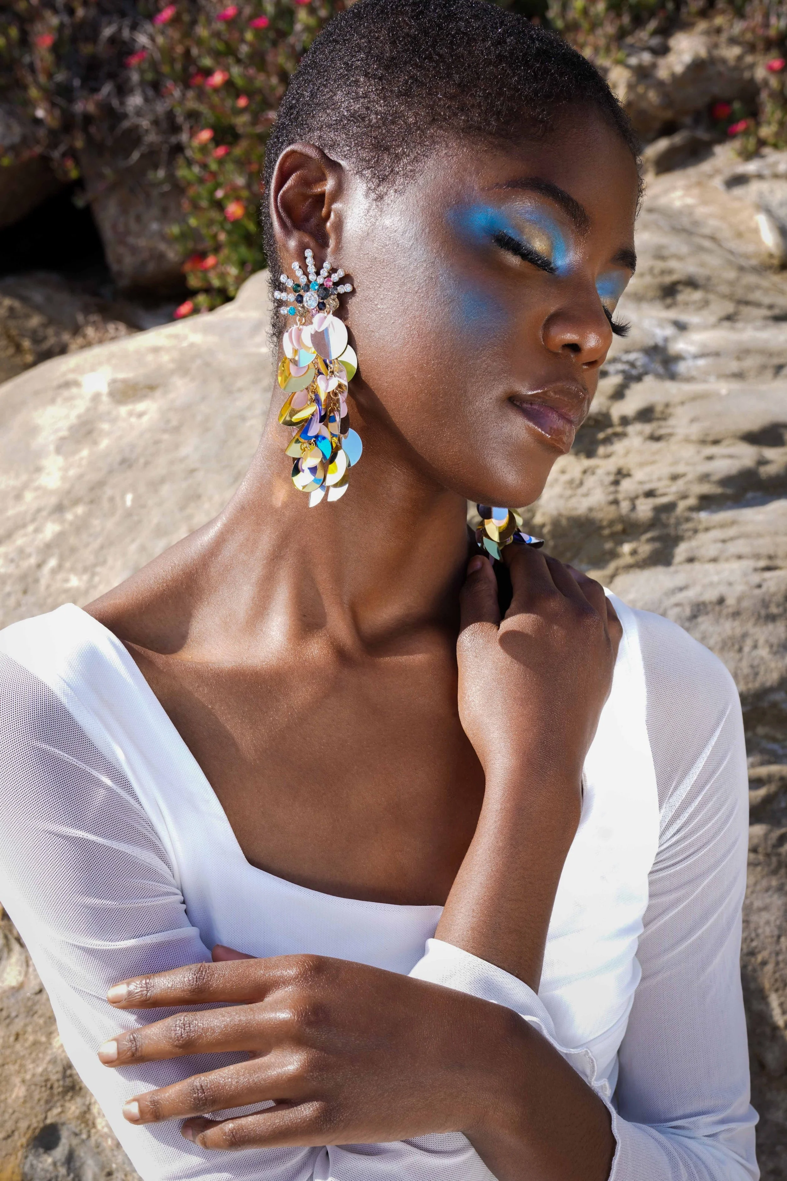 Black female model is posing in front of a brown rock with flowers. She is wearing blue makeup, a white dress and colorful jewelry. 