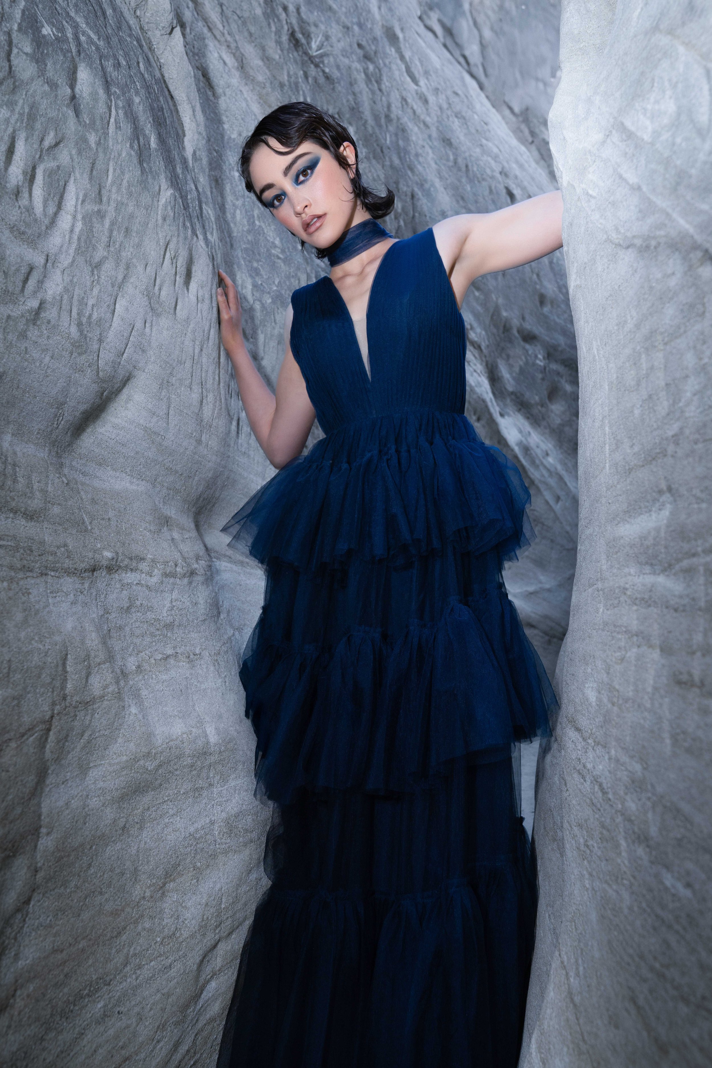 A fashion model wearing a flowing dark navy couture gown standing between dramatic canyon rock formations during a cinematic editorial fashion photoshoot. The low camera angle, movement in the sheer fabric, and confident expression create a powerful 
